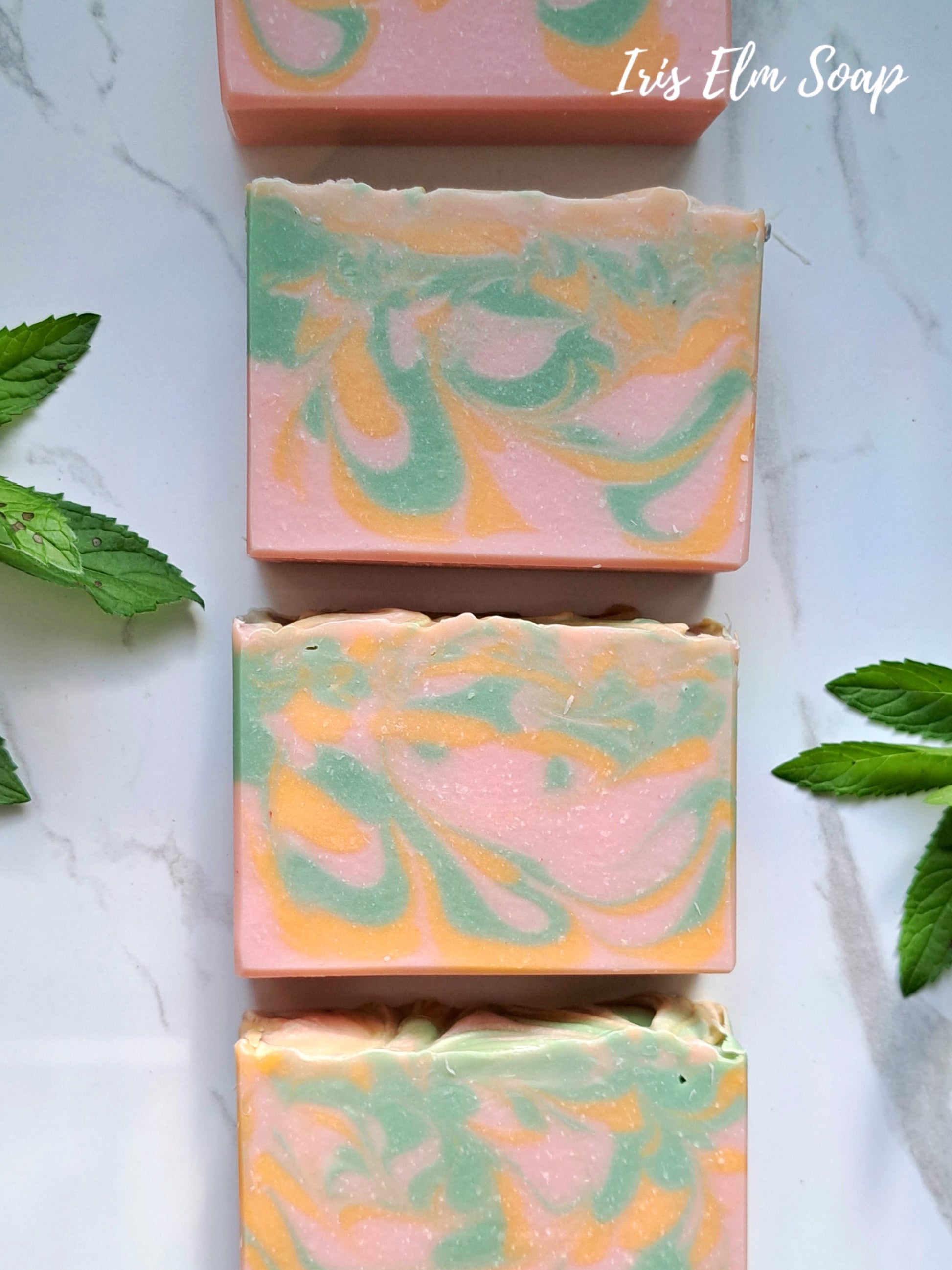 Swirled soap bars with pink, green, and orange colors on a white marble surface with 'Iris Elm Soap' branding.
