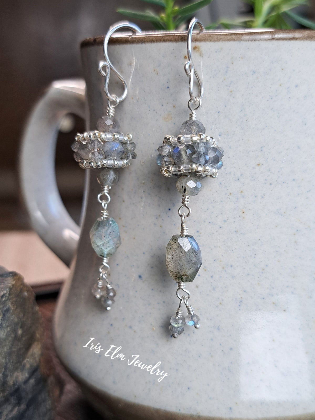 A pair of silver dangle earrings with faceted gray and iridescent labradorite beads, hanging from a speckled ceramic mug. The earrings feature silver hooks and beaded sections with dangling gray-green faceted beads and small clear crystal drops.