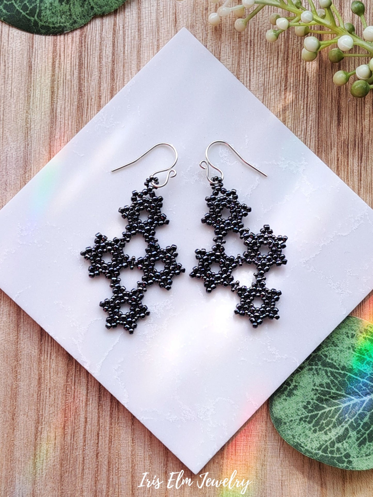beadwoven gothic dark pewter grey snowflake beaded earrings on white tile surrounded by greenery with Iris Elm Jewelry branding