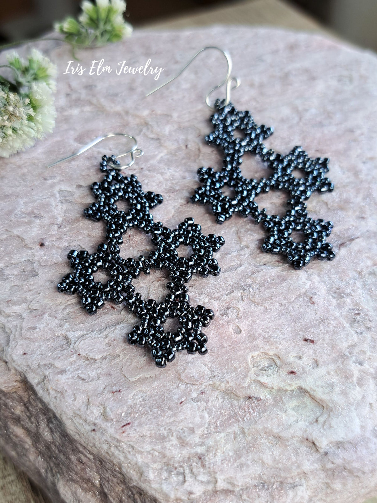 A pair of charcoal grey beaded snowflake dangle earrings with silver fishhook wires, displayed on a textured light pink stone surface with small green flowers in the corner. The brand name 'Iris Elm Jewelry' is watermarked in white text near the top left.