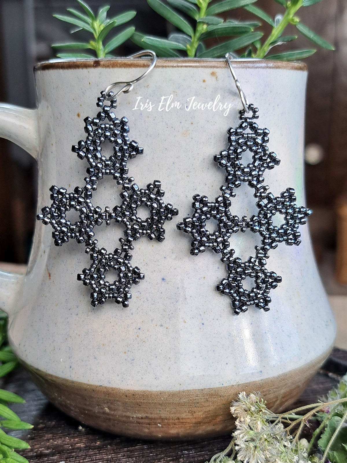 A pair of pewter grey beaded snowflake dangle earrings with silver fishhook wires, displayed on a speckled ceramic mug with green sprigs.