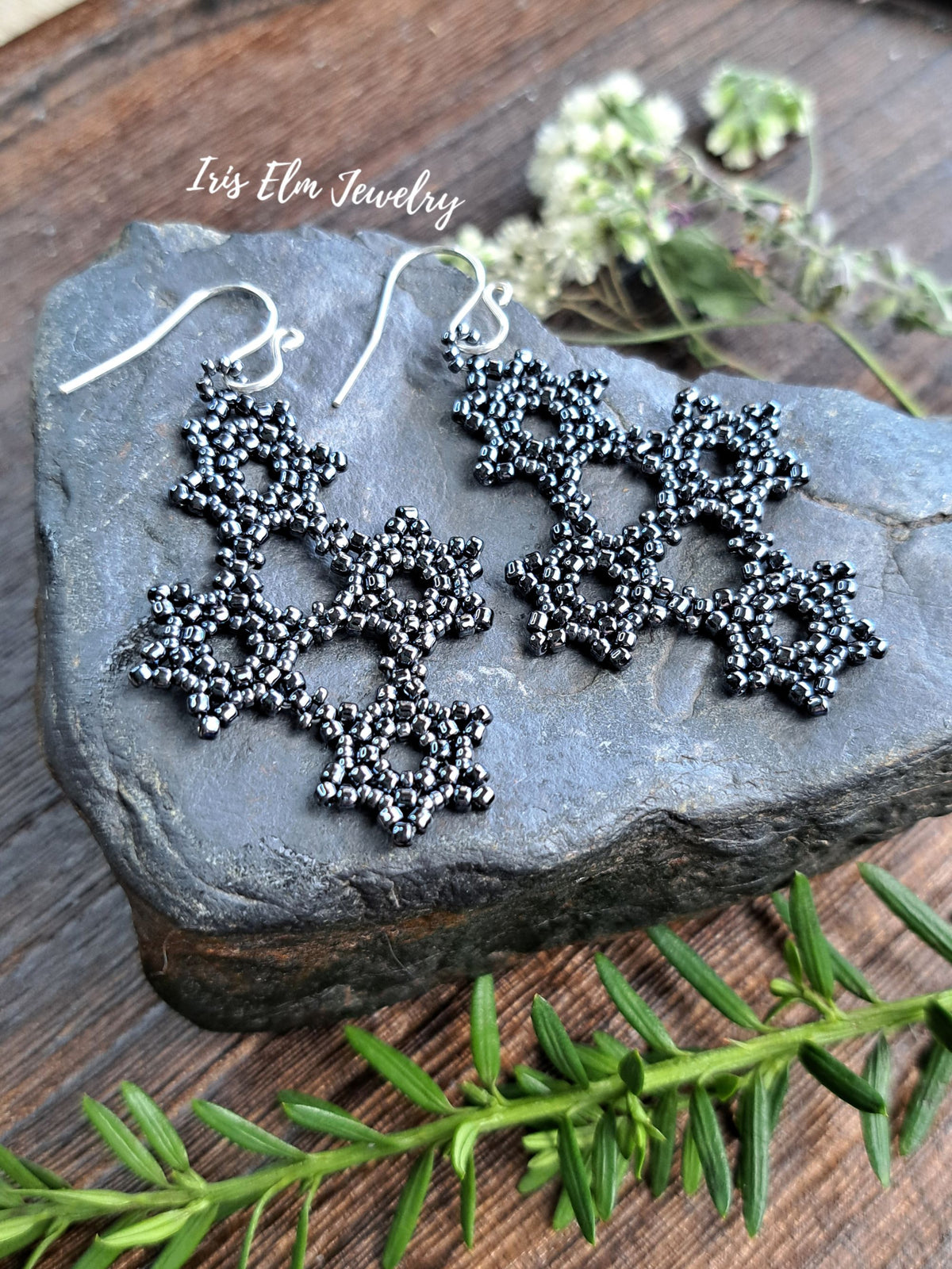 charcoal grey beaded snowflake dangle earrings displayed on a dark slate stone, with green sprigs and small white flowers on a rustic wooden surface. The brand name 'Iris Elm Jewelry' is watermarked near the top left.