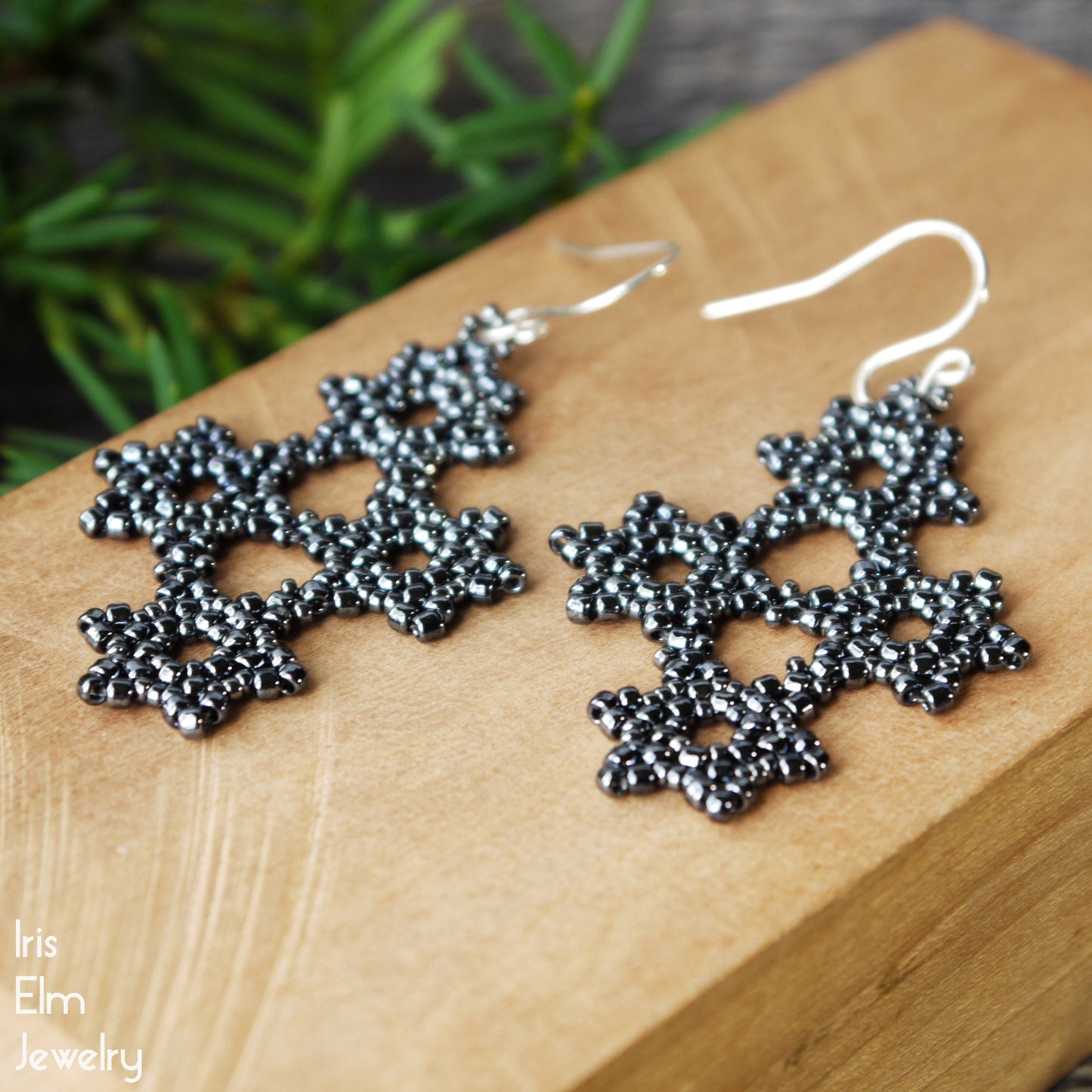 Dark grey snowflake earrings on a wooden block with a blurred green plant background