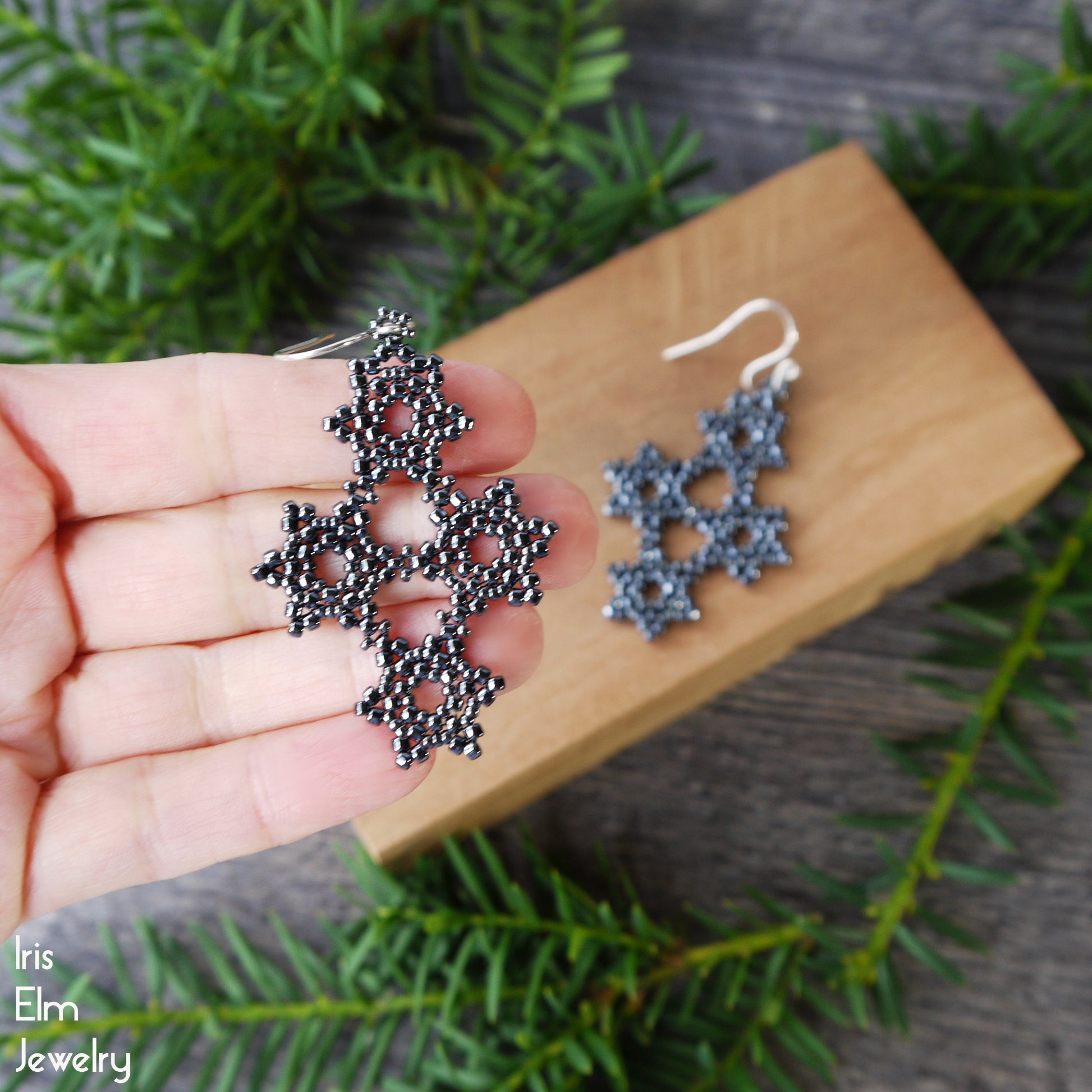Hand holding a pair of snowflake-shaped earrings with a wooden block and greenery in the background.