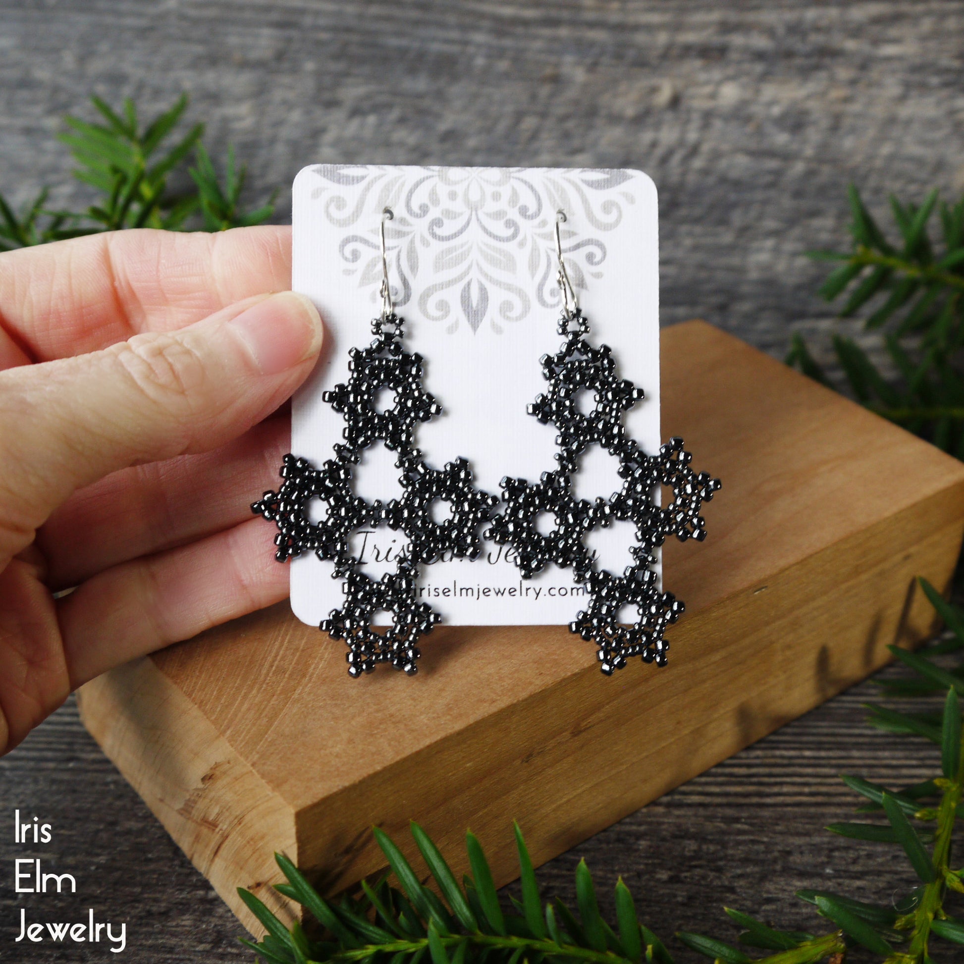 Dark charcoal grey beaded earrings on a card held by a hand with a wooden block and greenery in the background