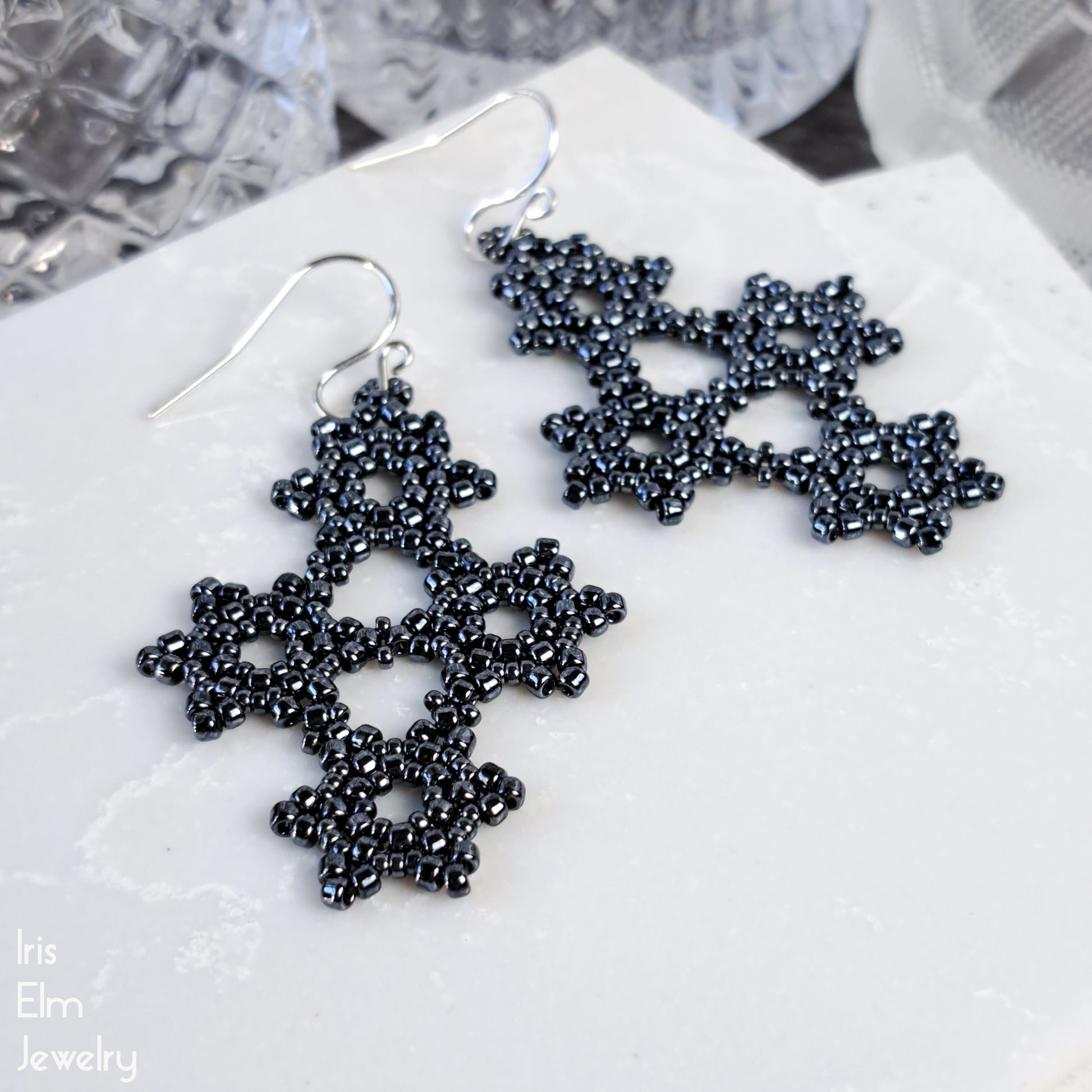 Dark grey woven beaded snowflake earrings on a white surface with 'Iris Elm Jewelry' branding.