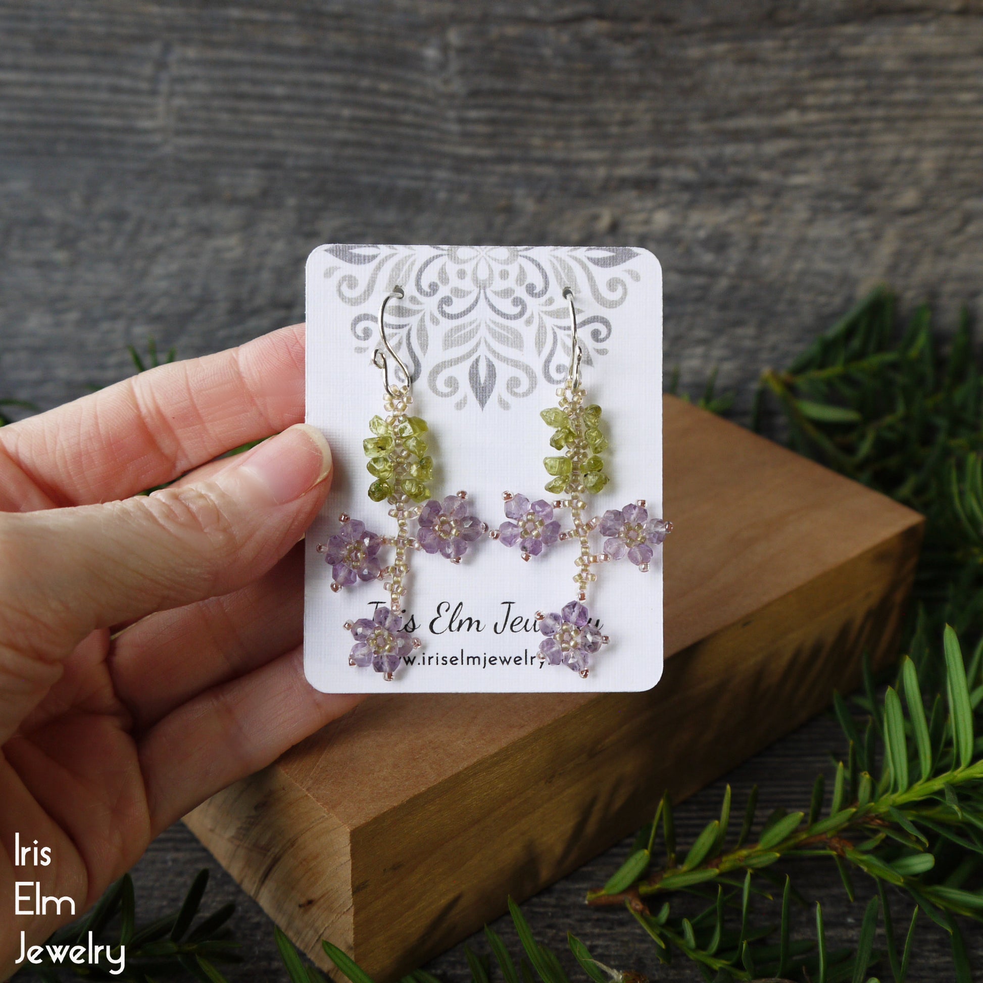 Pair of amethyst flower earrings on an 'Iris Elm Jewelry' card held by a hand with a natural background.
