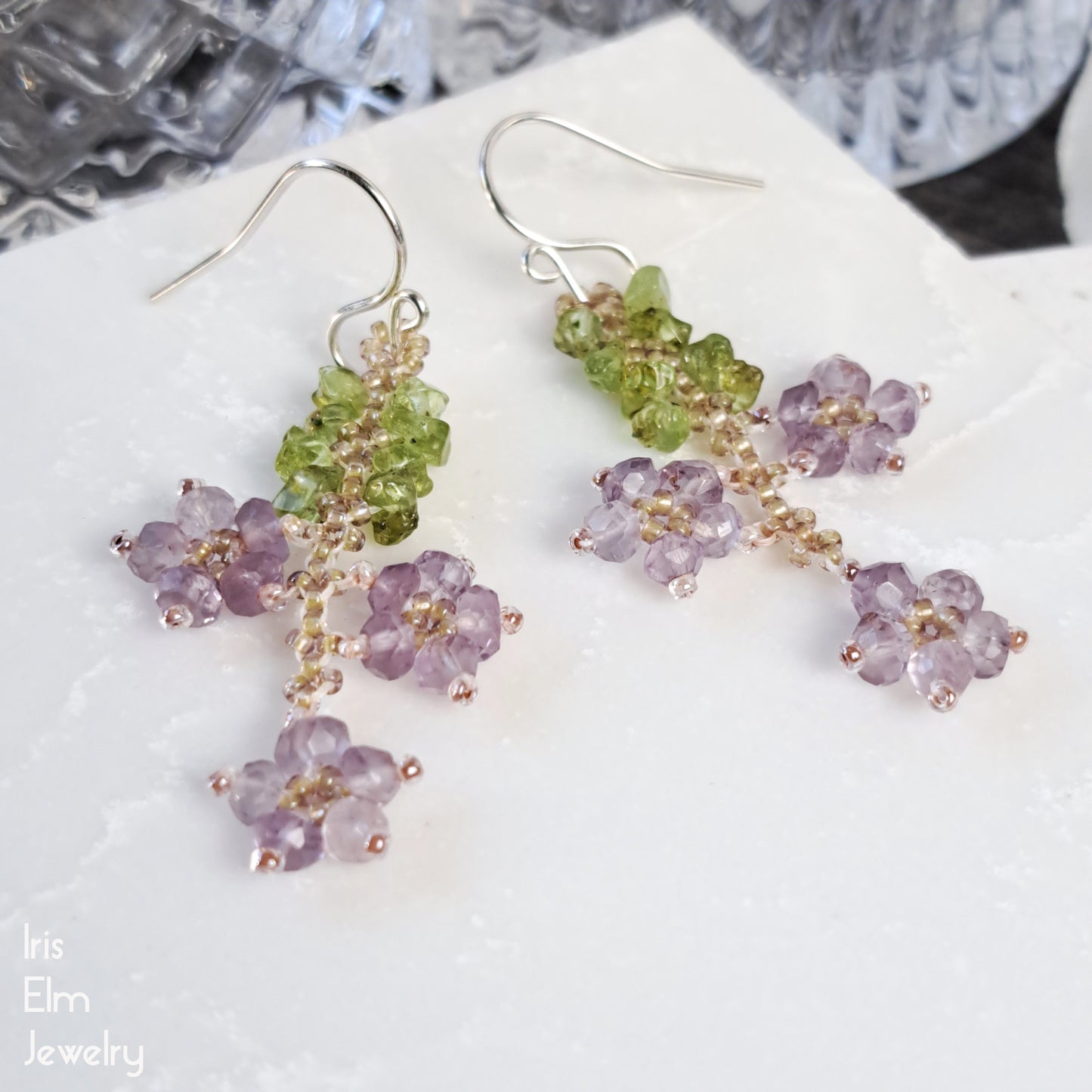 Pair of beaded flower earrings with green peridot and purple amethyst gemstones on a white surface.