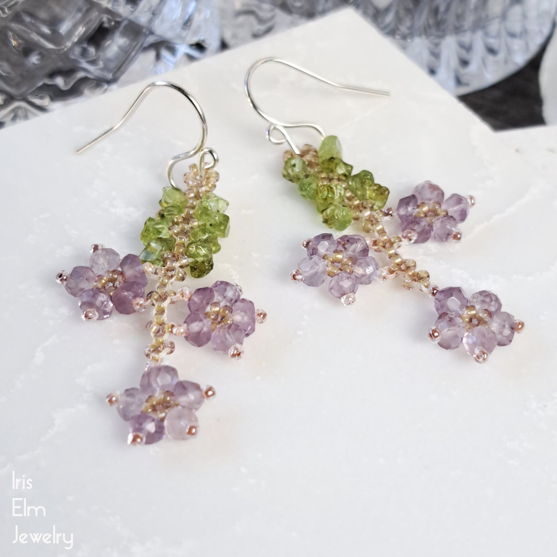 Pair of amethyst beadwork floral earrings with green and purple stones on a white surface.