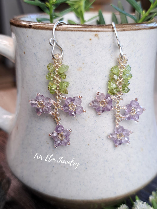 A pair of handmade dangle earrings by Iris Elm Jewelry, featuring light green peridot beads and lavender amethyst flower clusters on silver hooks, displayed on a speckled ceramic mug.