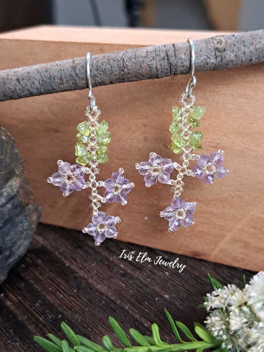A pair of handmade silver dangle earrings with green and lavender gemstone flower and leaf beadwork clusters, displayed on a rustic wood branch with a brown wooden block background.