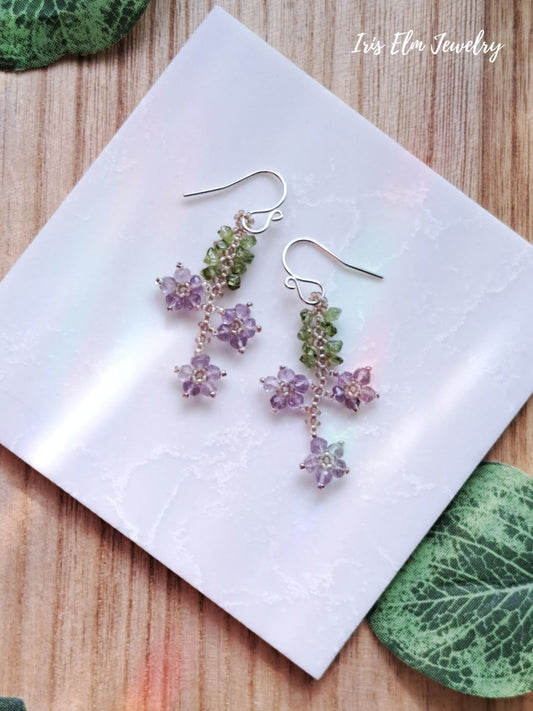 Pair of floral beadwork amethyst and peridot flower earrings on a white card with 'Iris Elm Jewelry' branding.