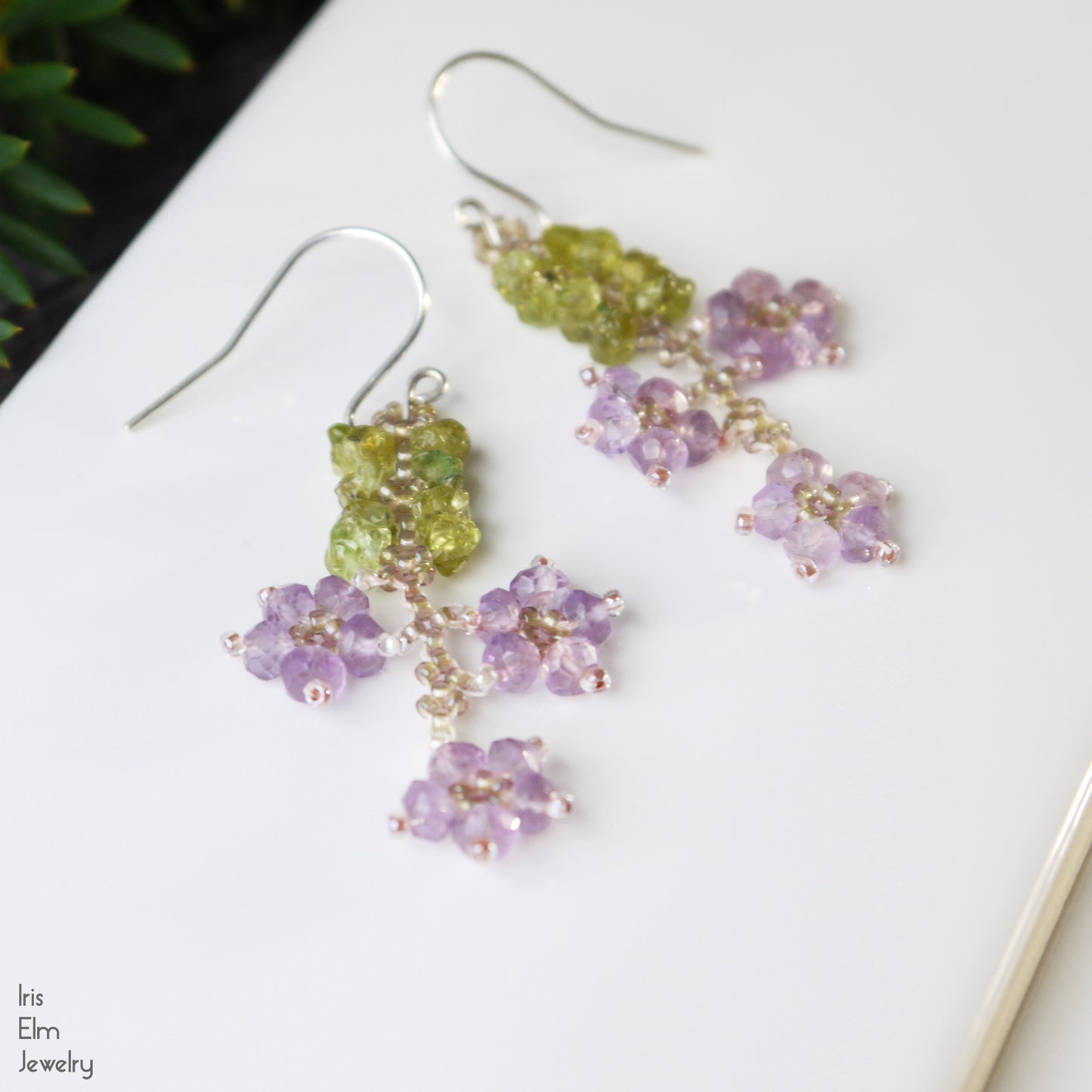 Pair of green and purple beaded flower earrings on a white surface with 'Iris Elm Jewelry' branding.