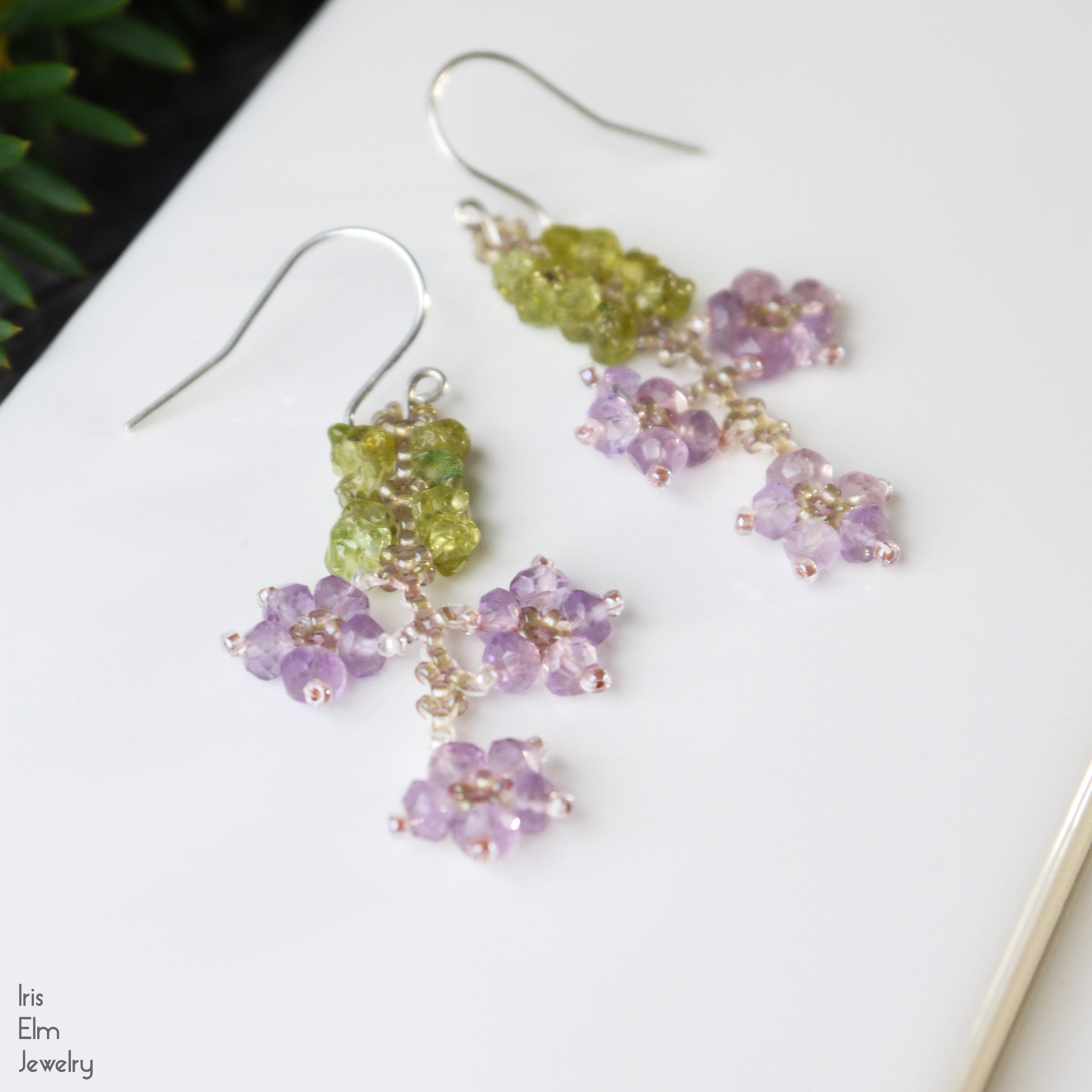 Pair of green and purple beaded flower earrings on a white surface with 'Iris Elm Jewelry' branding.
