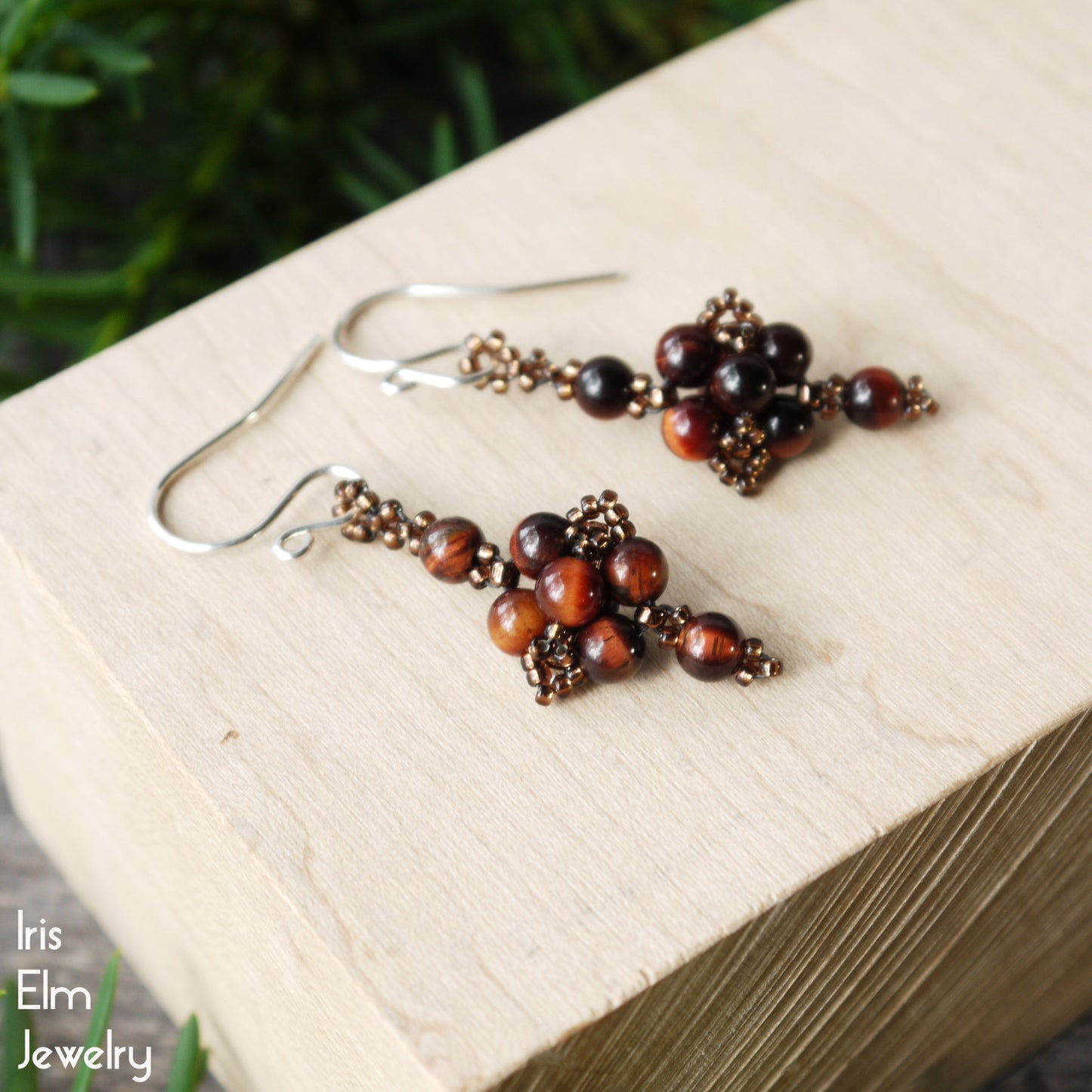 Pair of beaded handmade red tiger eye earrings on a wooden block with 'Iris Elm Jewelry' branding.