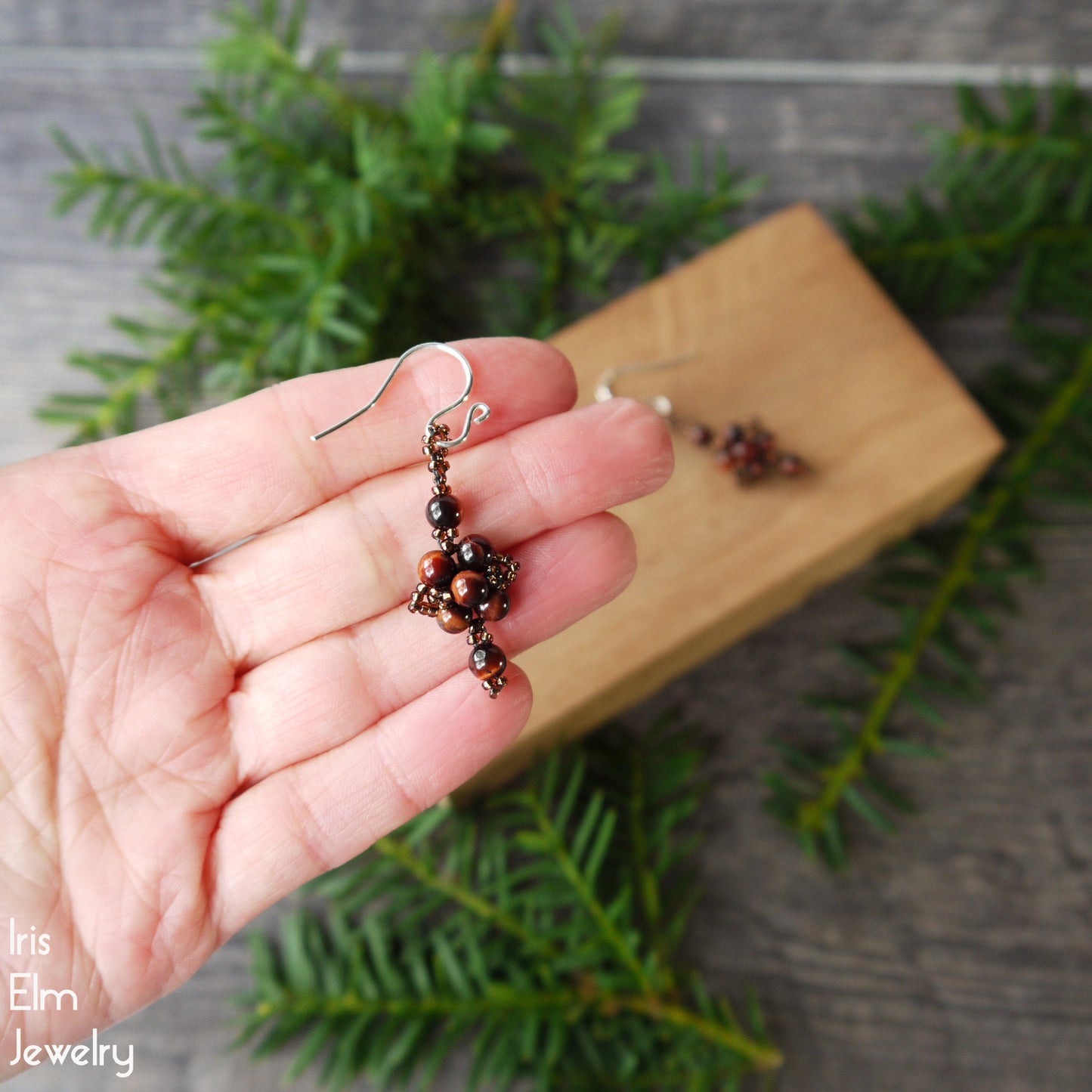 Hand holding a beaded tigers eye earring with a wooden block and greenery in the background