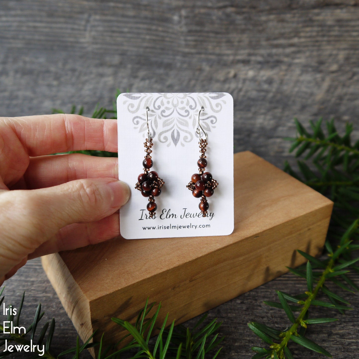 Pair of beaded brown stone earrings on a 'Iris Elm Jewelry' card held by a hand with a wooden block and greenery in the background.