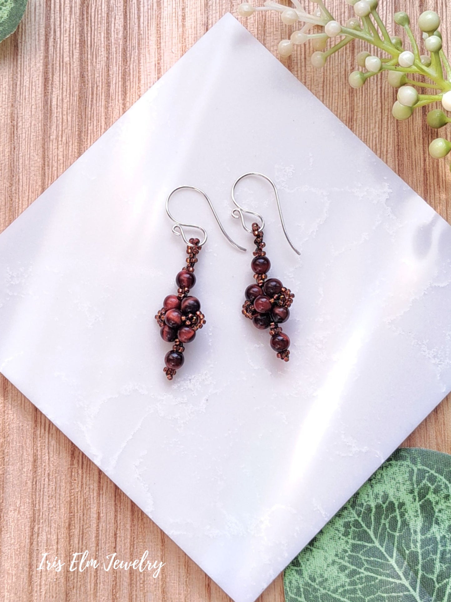 Pair of red tiger eye small beaded earrings on a white tile with green leaves in the background