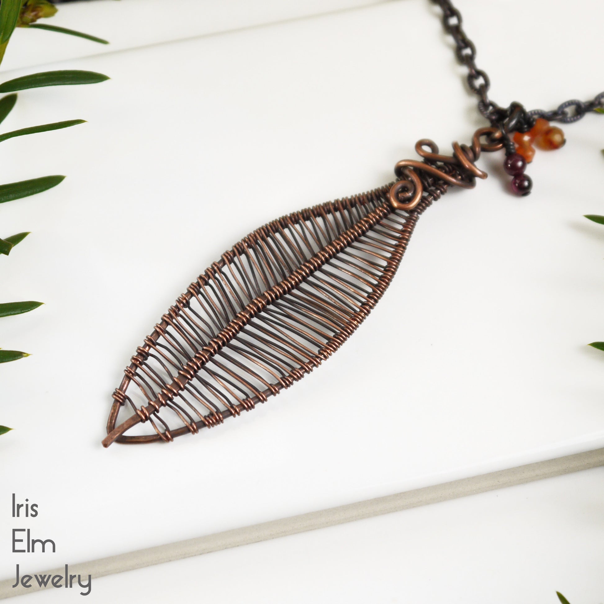 Copper leaf-shaped necklace with orange carnelian and red garnet beads on a white background, featuring 'Iris Elm Jewelry' branding.