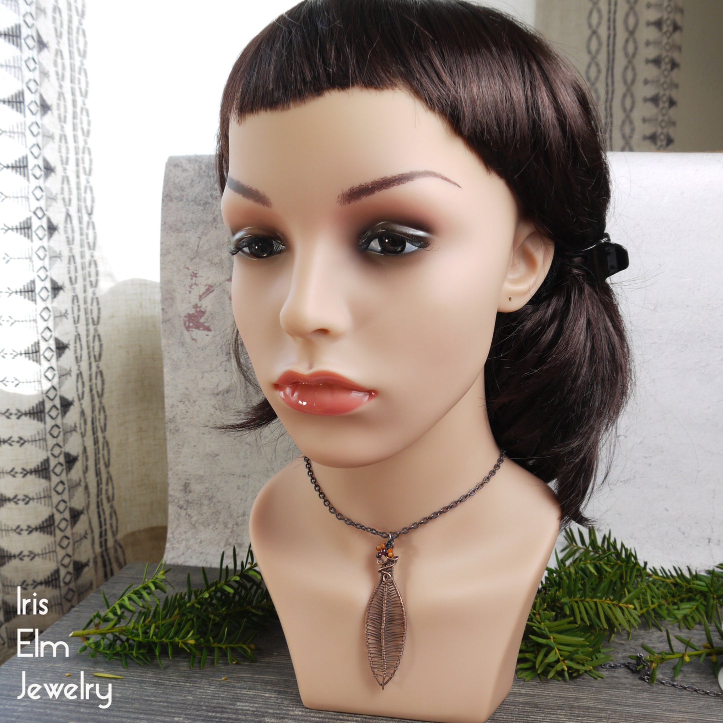 Mannequin head with brown hair and a leaf pendant necklace on a decorative surface with 'Iris Elm Jewelry' branding.