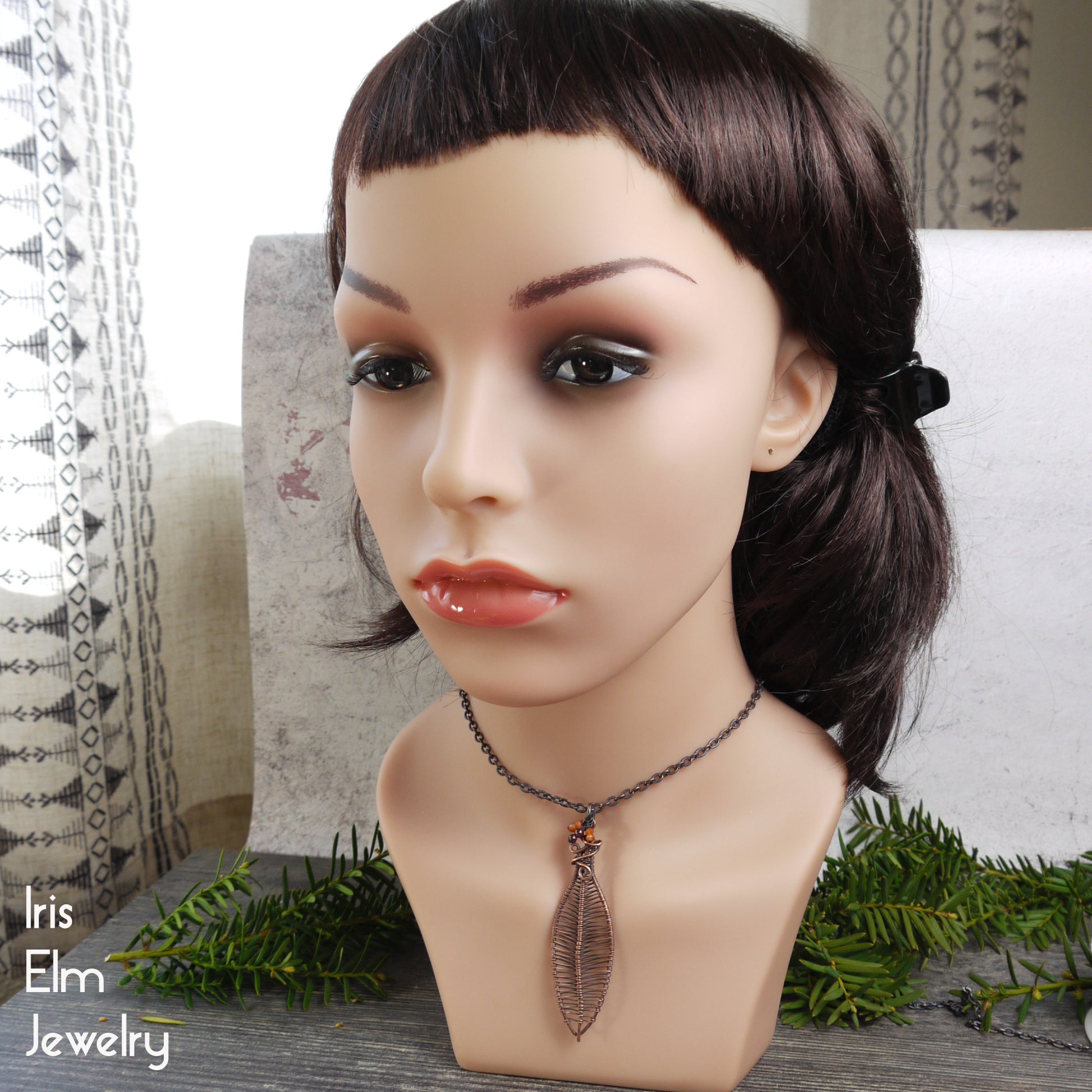 Mannequin head with brown hair and a leaf pendant necklace on a decorative surface with 'Iris Elm Jewelry' branding.