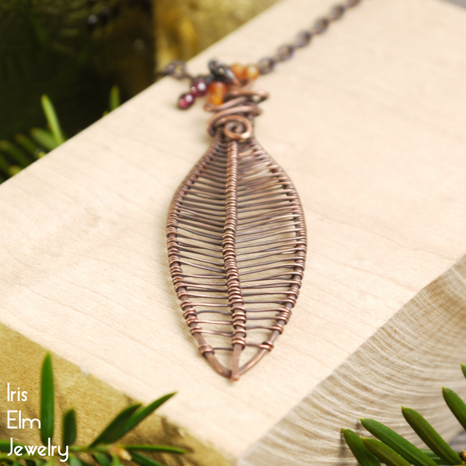 Copper leaf-shaped pendant necklace on a wooden surface with greenery in the background