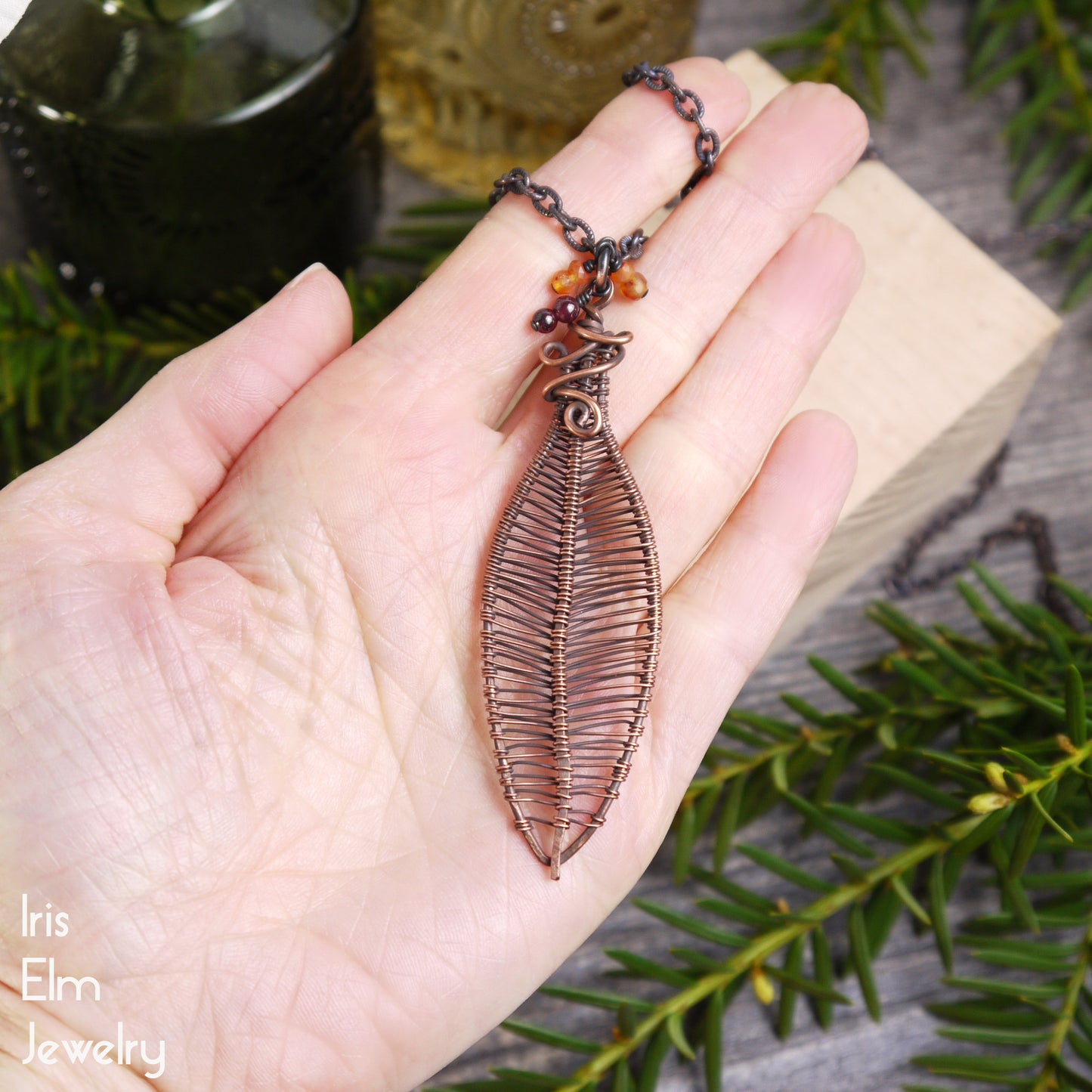 Hand holding a leaf-shaped copper wire wrapped pendant with a chain, surrounded by greenery.