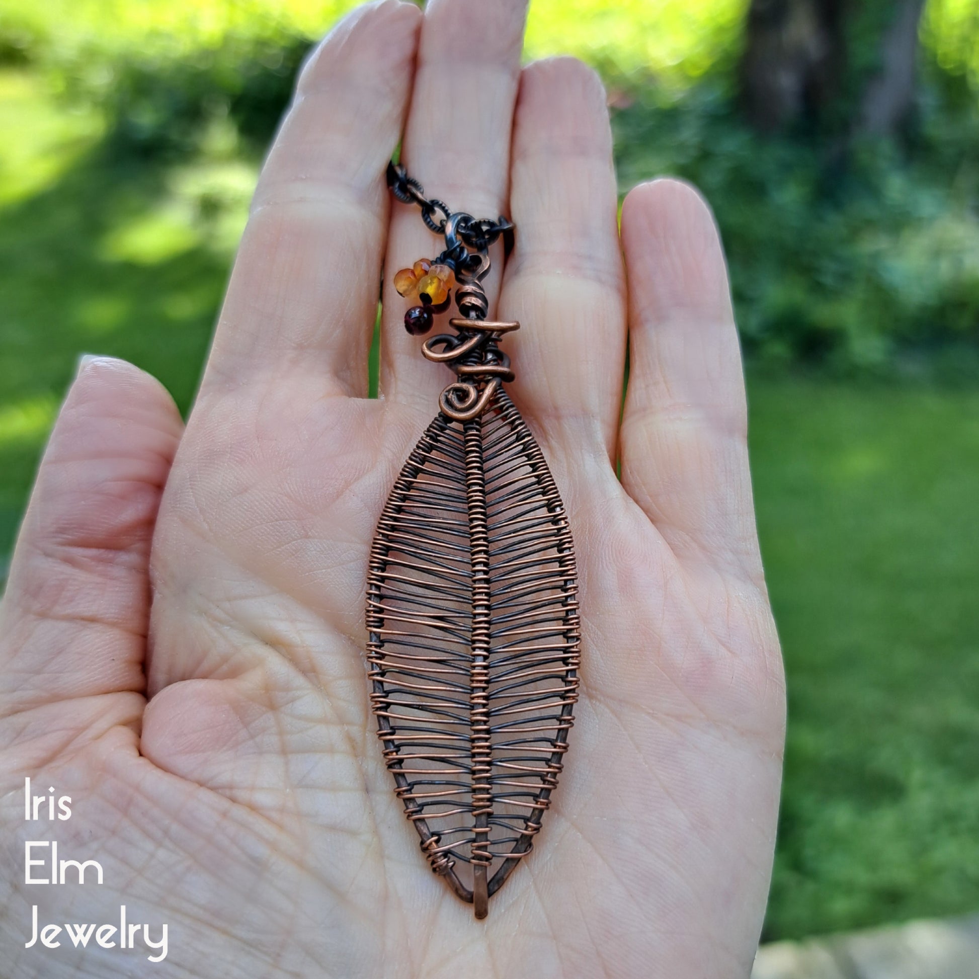 Hand holding a leaf-shaped copper wire woven pendant with a blurred green outdoor background