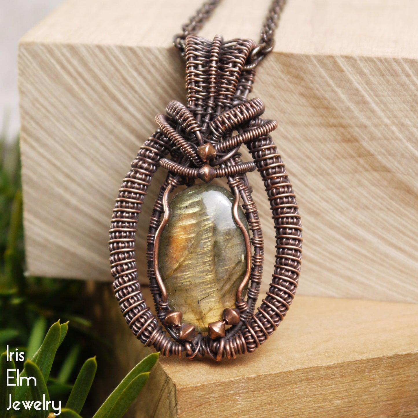 copper coil wire wrapped handmade Jewelry pendant with a labradorite stone on a wooden block, brand 'Iris Elm Jewelry' visible