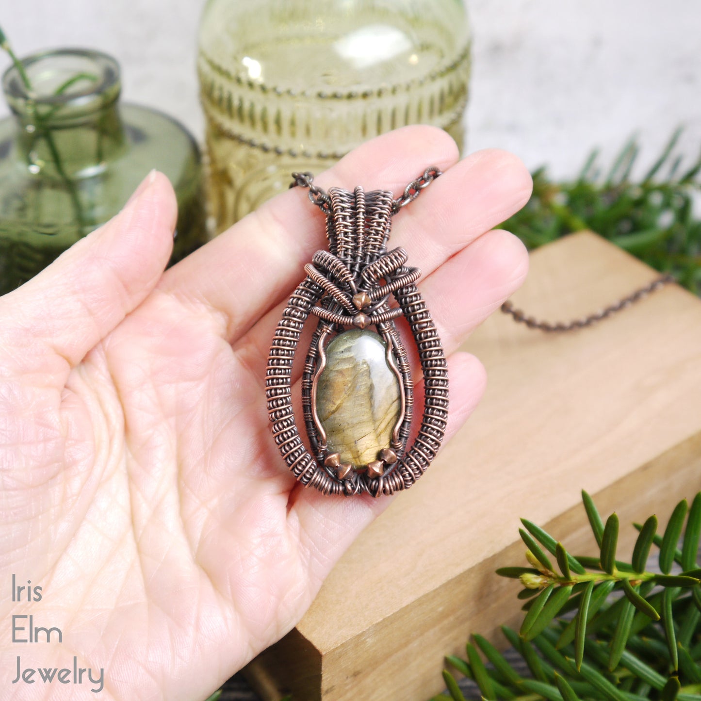 Hand holding a decorative copper wire woven labradorite pendant with a stone, set against a natural background with plants and jars