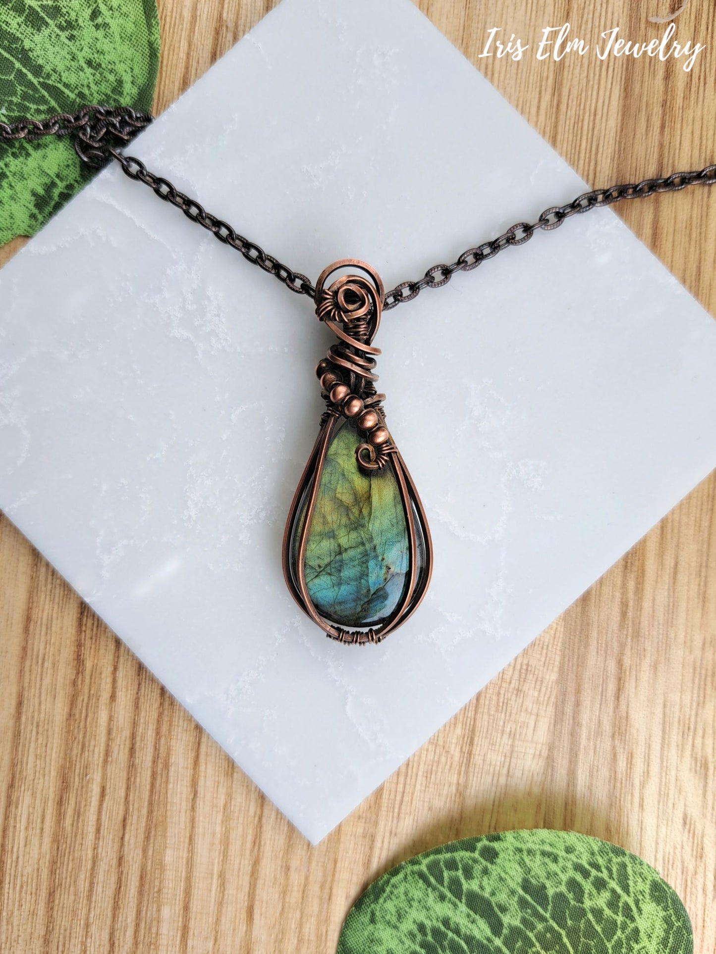 A handmade teardrop pendant necklace featuring a marbled green and blue labradorite stone wrapped in copper wire, hanging from a dark metal chain on a white textured card over a wooden surface.