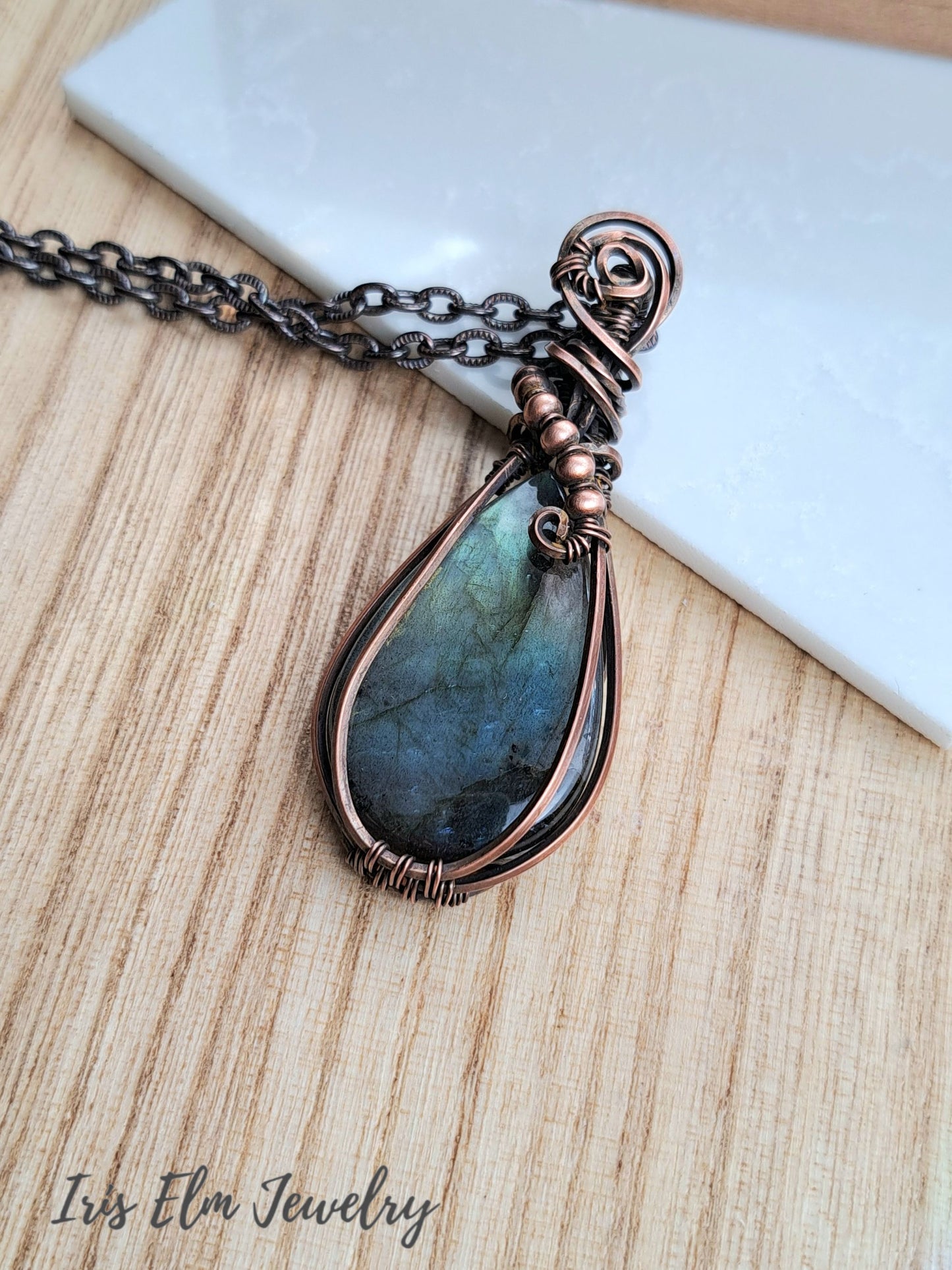A teardrop-shaped labradorite pendant necklace wrapped in copper wire, resting on a light wood surface next to a white marble tile. The brand name 'Iris Elm Jewelry' is visible in the bottom-left corner.