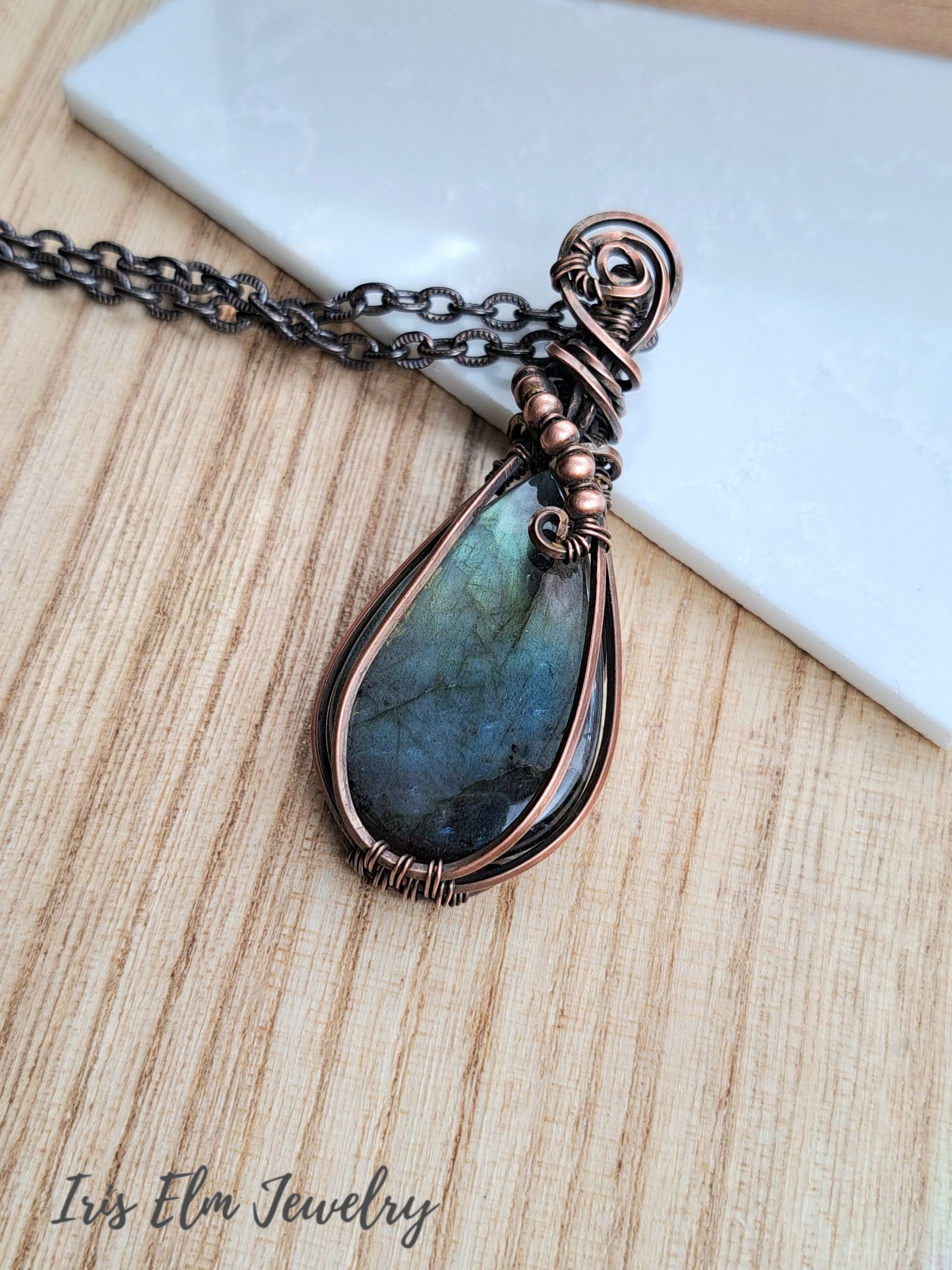 A teardrop-shaped labradorite pendant necklace wrapped in copper wire, resting on a light wood surface next to a white marble tile. The brand name 'Iris Elm Jewelry' is visible in the bottom-left corner.