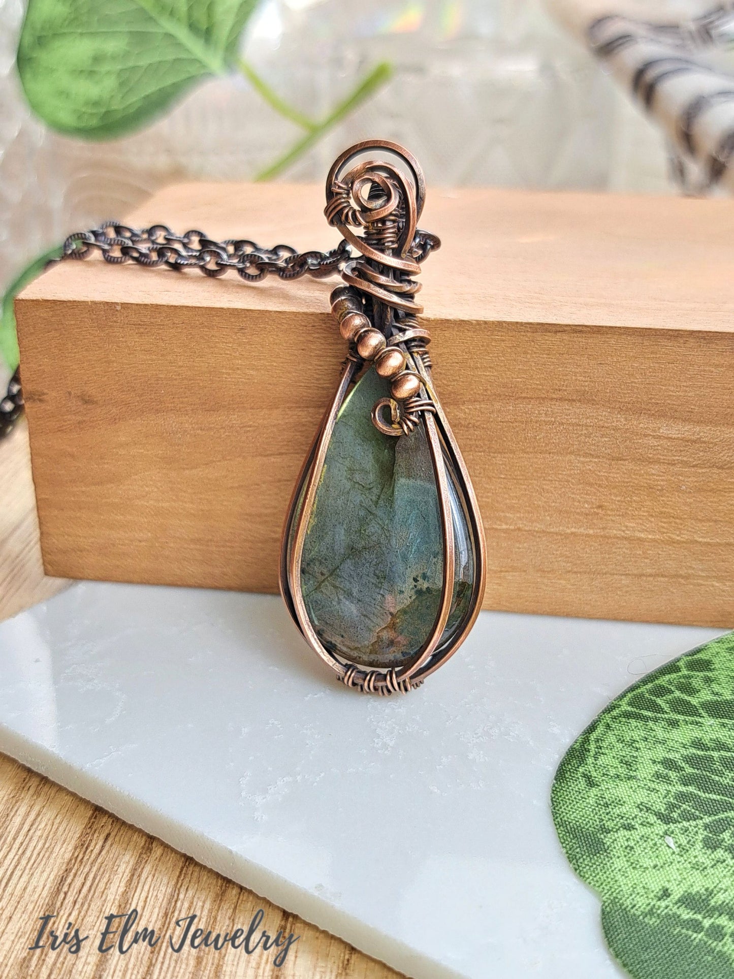 A handmade teardrop pendant necklace featuring a greenish-blue stone wrapped in copper-toned metal, resting on a wooden block with a white marble surface below. The brand name 'Iris Elm Jewelry' is visible in the bottom-left corner.