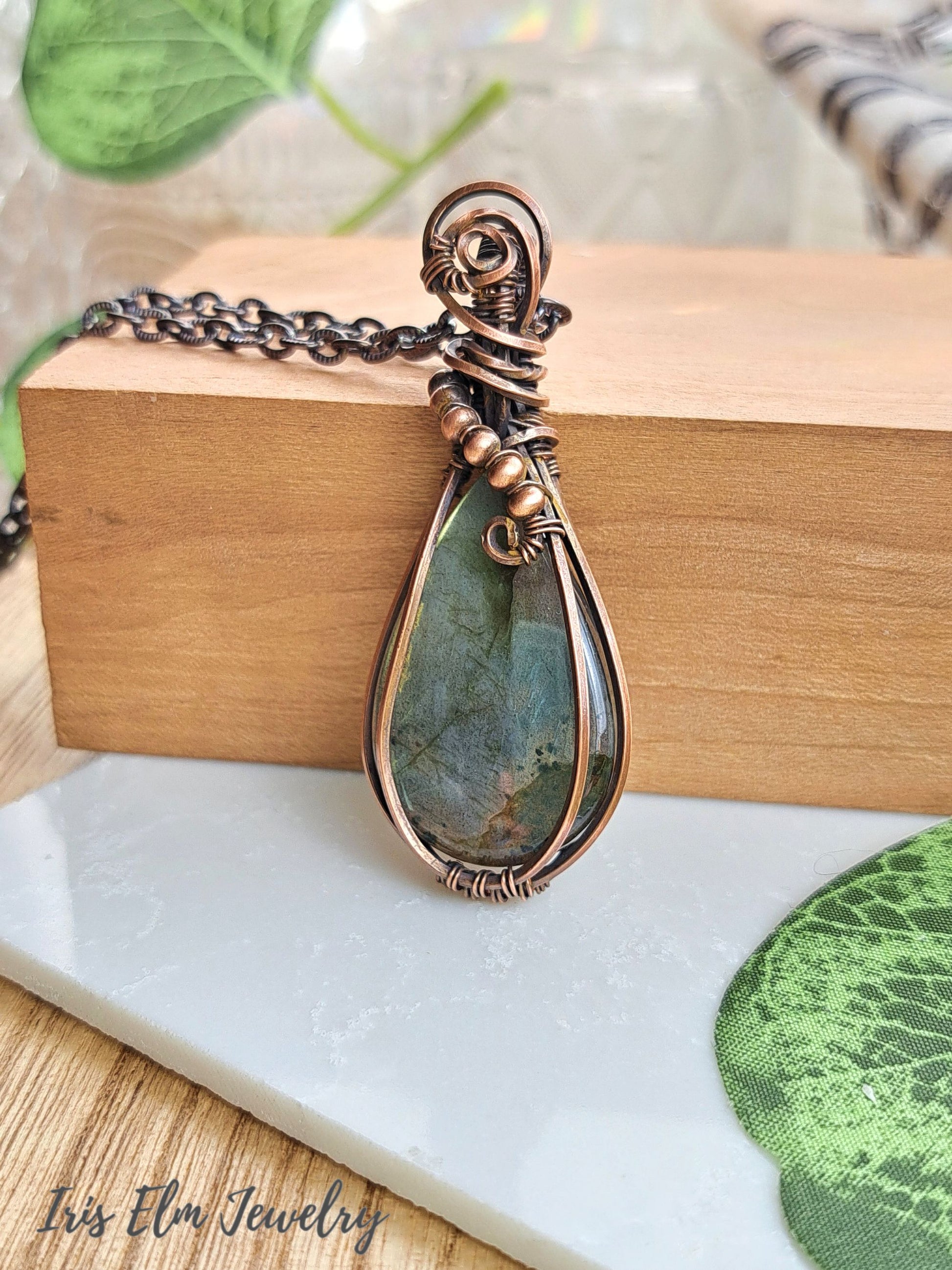 A handmade teardrop pendant necklace featuring a greenish-blue stone wrapped in copper-toned metal, resting on a wooden block with a white marble surface below. The brand name 'Iris Elm Jewelry' is visible in the bottom-left corner.