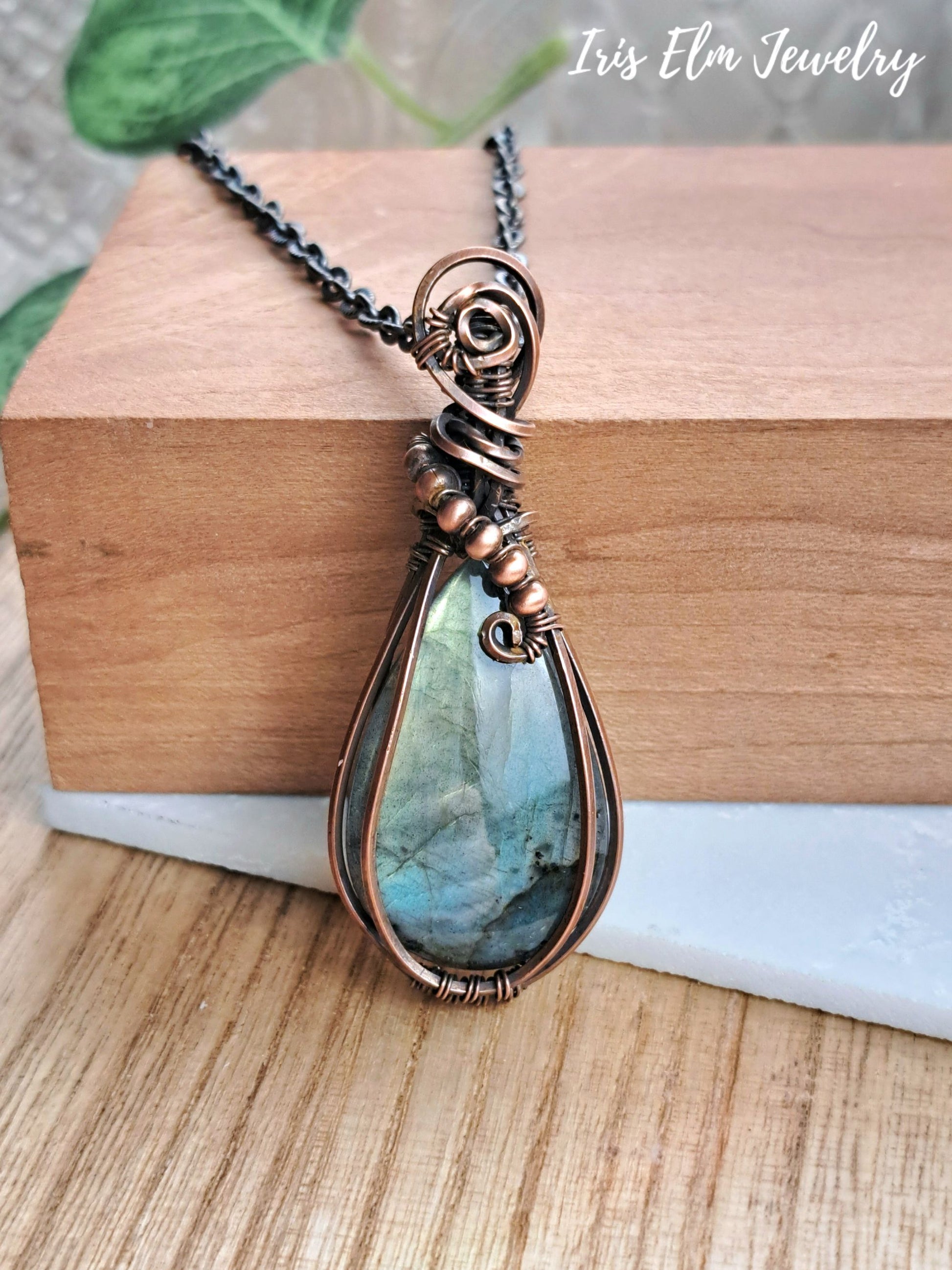 A teardrop-shaped pendant necklace with a blue-green labradorite stone wrapped in copper-toned metal, resting on a wooden block with a copper chain.