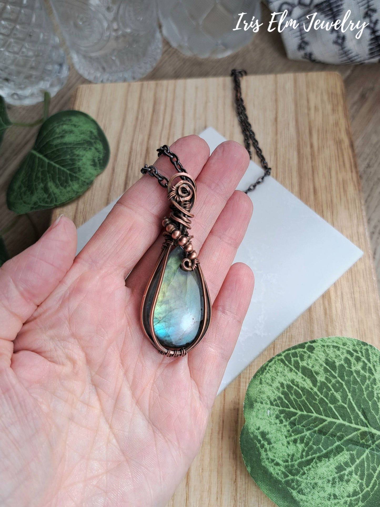A hand holds a teardrop-shaped pendant with a shimmering iridescent blue-green labradorite gemstone and dark copper-toned metal wrapping, shown against a wooden surface with decorative leaves and glassware. The text 'Iris Elm Jewelry' appears in the top right.