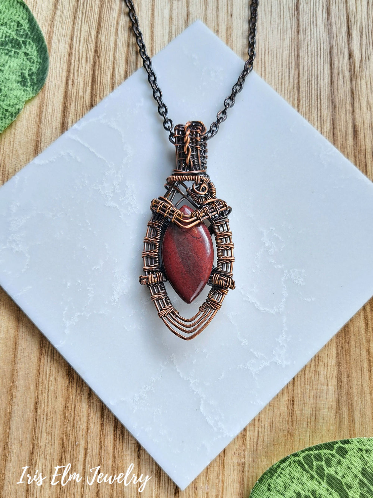 A handmade pendant necklace with a copper wire-wrapped setting holding a red jasper stone, displayed on a white textured square tile over a wooden surface.