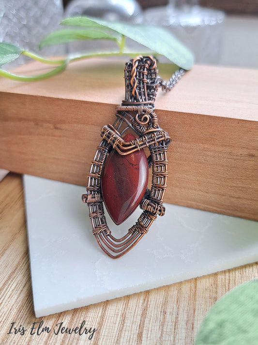 A handmade copper wire-wrapped pendant necklace featuring a polished dark red marquise-shaped red jasper stone, displayed on a wooden block with a white marble tile underneath. The brand name 'Iris Elm Jewelry' is visible in the bottom-left corner.