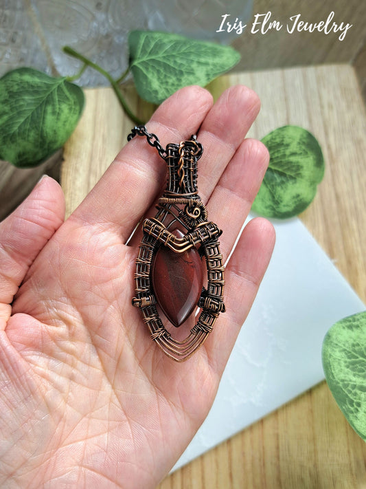 A hand holding a handmade pendant necklace with a dark reddish-brown teardrop red jasper stone set in an ornate bronze-toned metal cage, with the brand name 'Iris Elm Jewelry' visible in the top right corner.