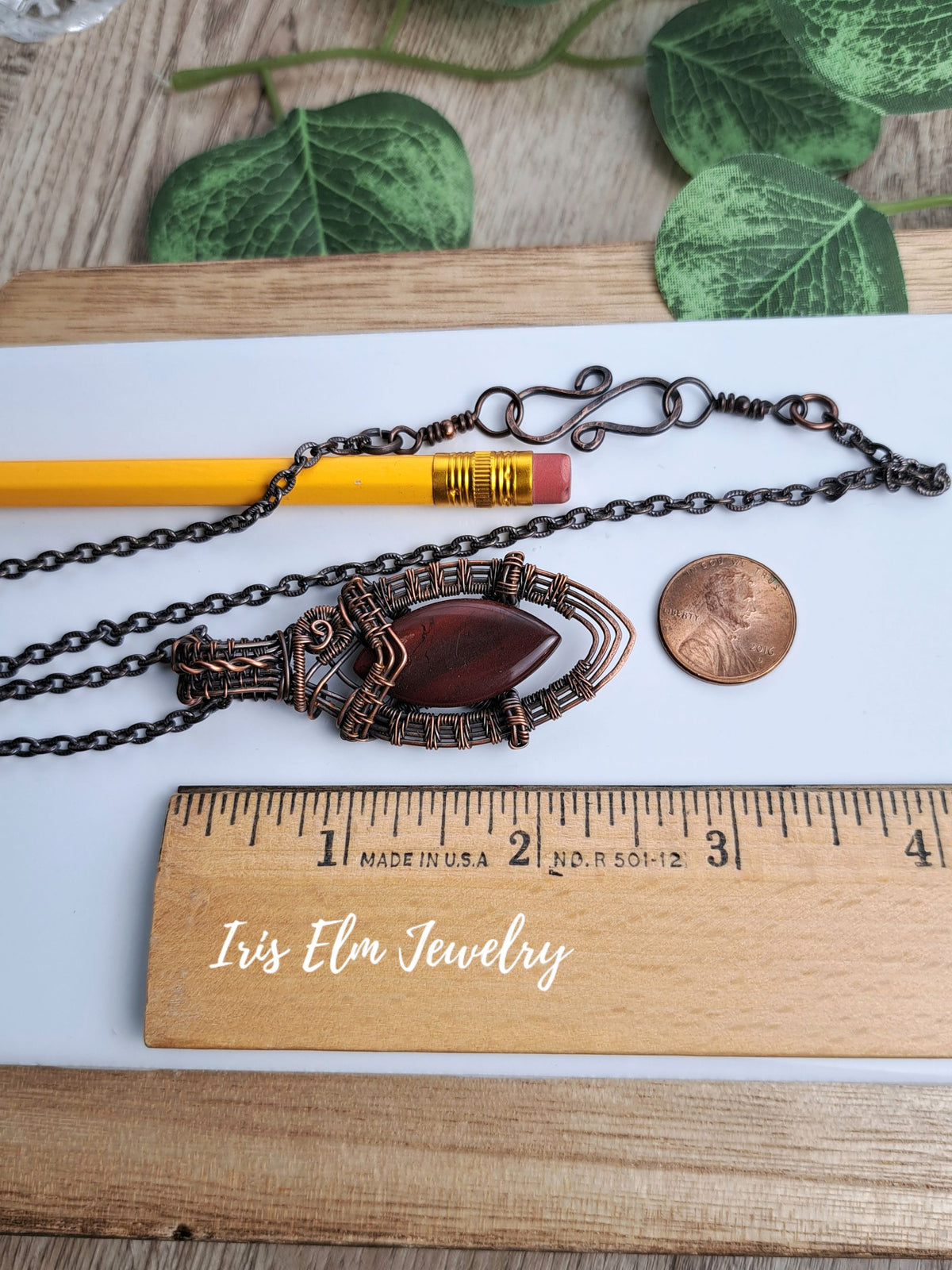 A handmade 2.5 inch long pendant necklace with a dark bronze-toned metal frame and a reddish-brown stone, displayed on a white sheet with a yellow pencil, penny, wooden ruler, and green leaves in the background. The text 'Iris Elm Jewelry' is written on the ruler.