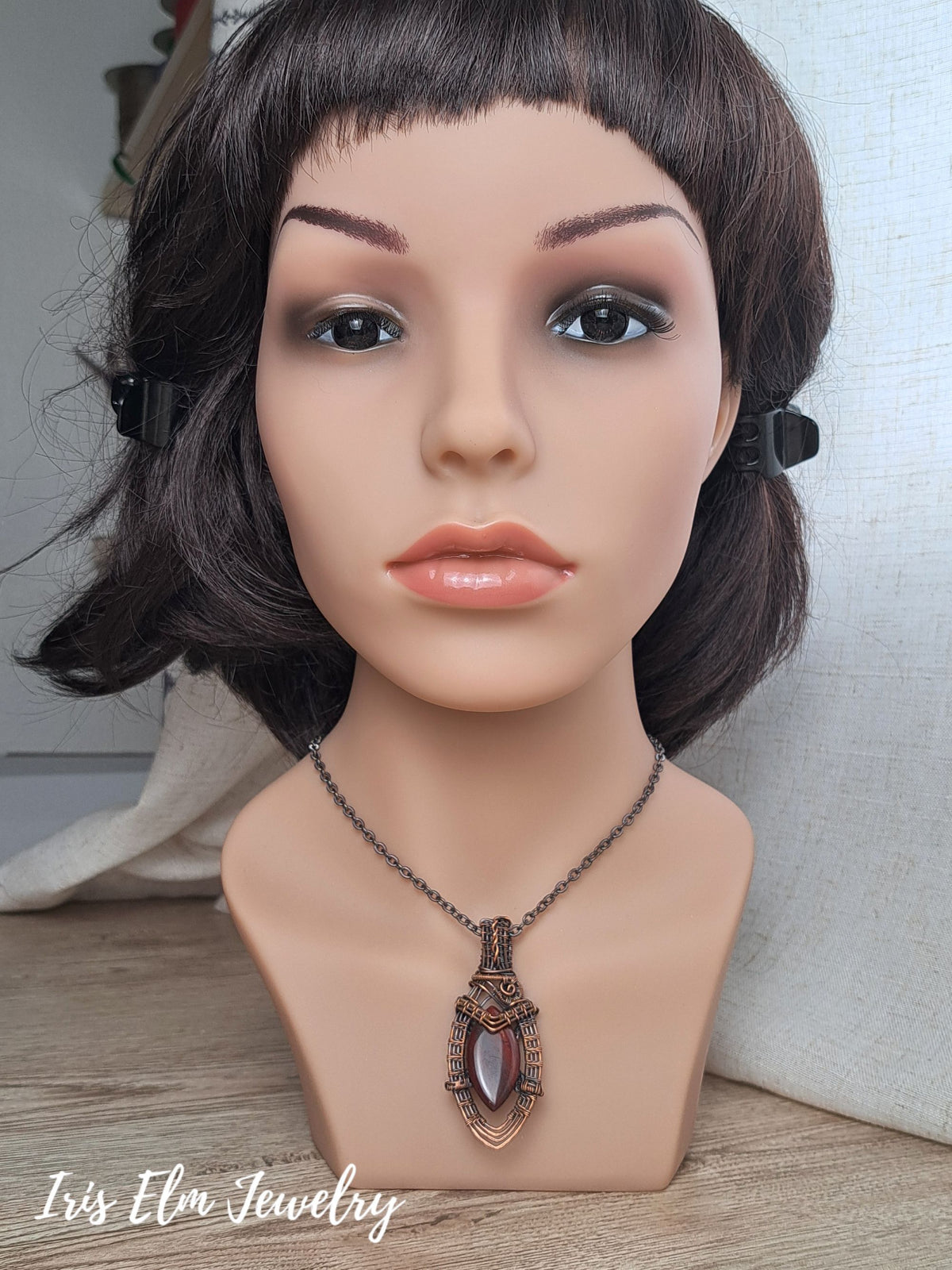 A close-up of a mannequin bust wearing a dark chain copper necklace with an ornate bronze-toned pendant featuring a central red jasper stone. The mannequin has short dark brown hair and is photographed indoors against a light neutral backdrop, with the text 'Iris Elm Jewelry' visible in the lower left.