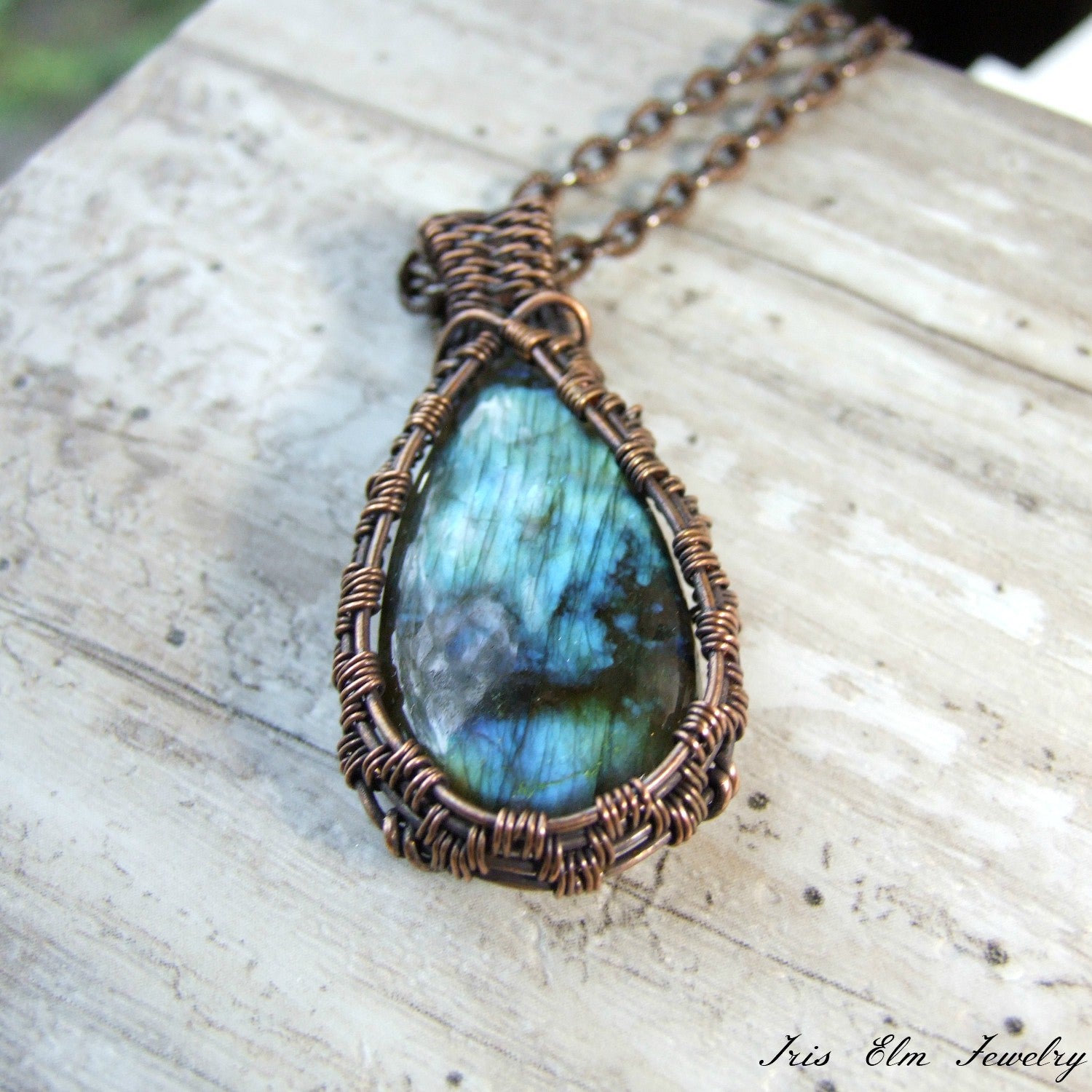 Teardrop-shaped labradorite pendant in a copper wire wrap setting on a light wooden surface.