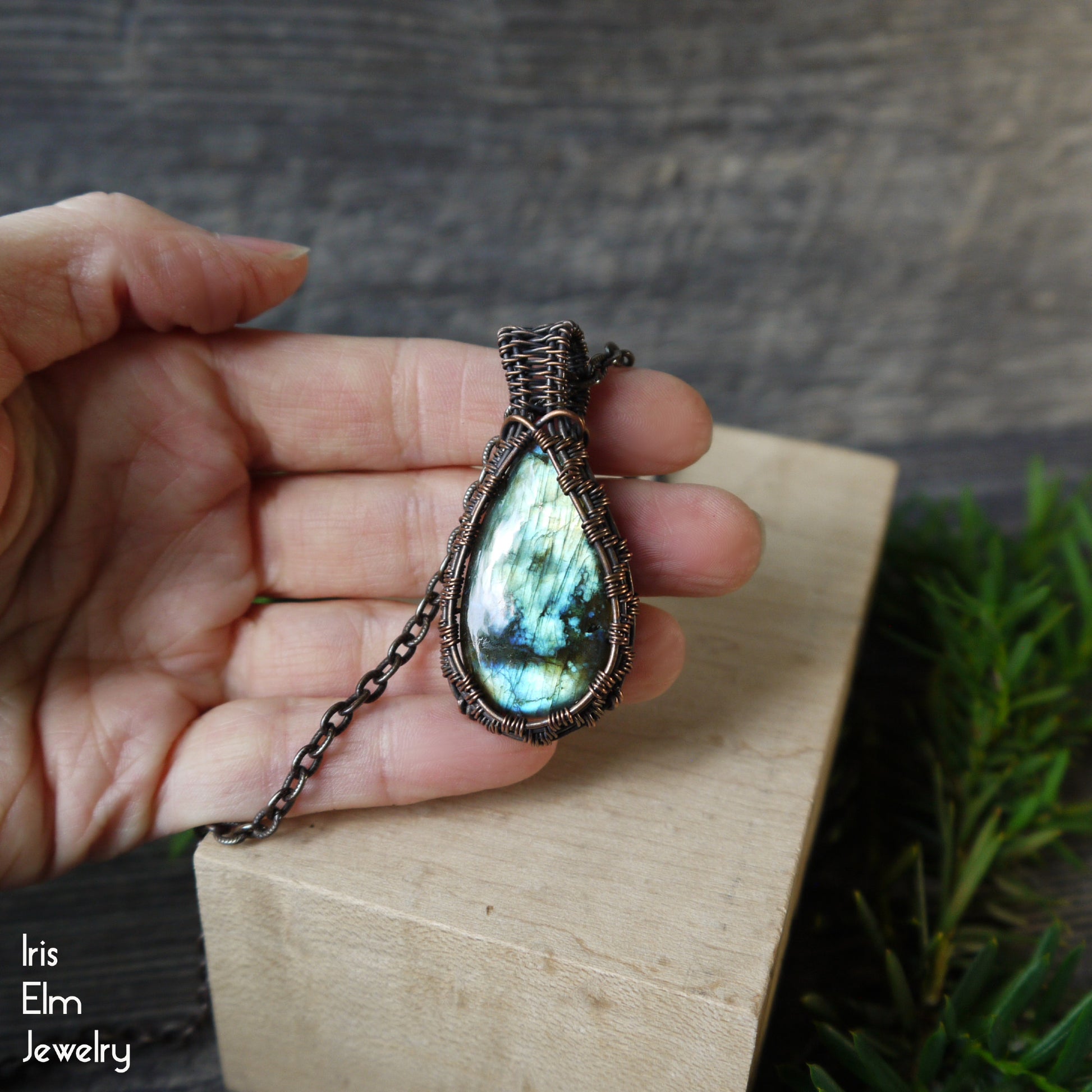Hand holding a copper wire wrapped Labradorite pendant necklace with a wooden block and blurred greenery in the background
