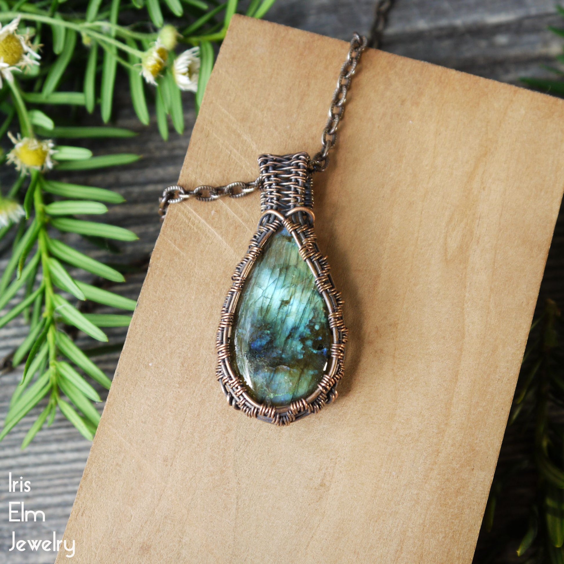 Teardrop-shaped oxidized copper handmade pendant with a green iridescent labradorite gemstone on a wooden surface, surrounded by greenery.