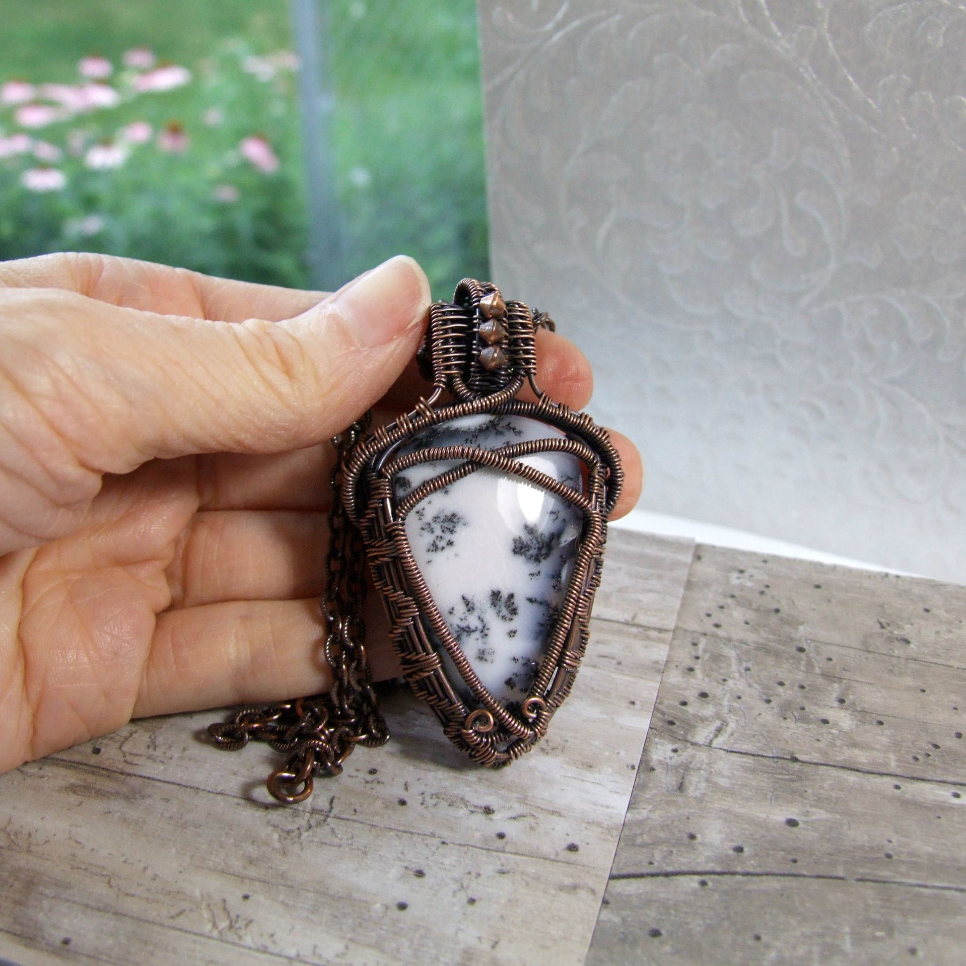 Hand holding a copper-wrapped white dendritic agate stone pendant on a wooden surface with a blurred outdoor background