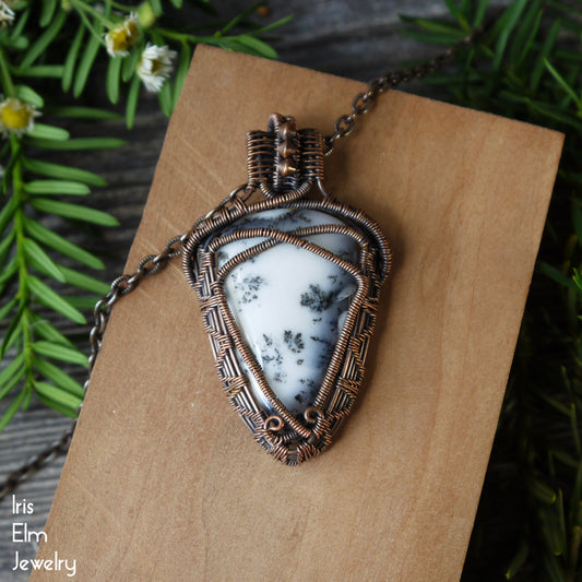 Jewelry pendant with white dendritic agate stone and oxidized copper wire wrapping on a wooden surface, surrounded by greenery.