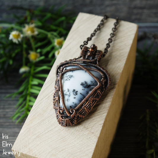 Necklace with a dendritic agate triangle stone pendant that is wrapped in oxidized copper wire weaving on a wooden block, surrounded by greenery.