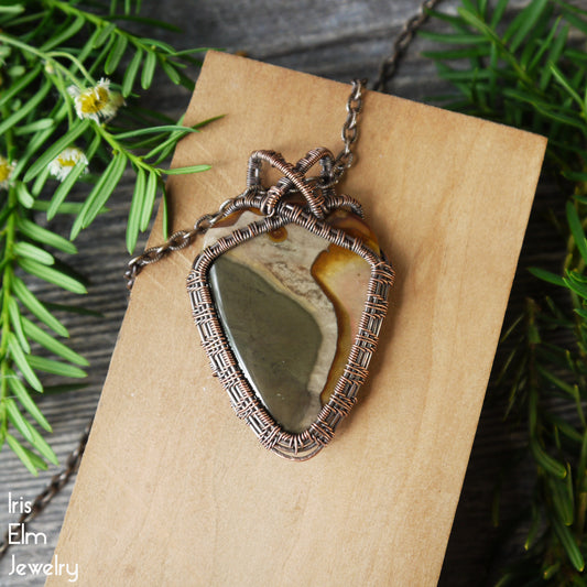 Necklace with a oxidized copper wire-wrapped inverted triangle impression jasper pendant on a wooden block with greenery in the background