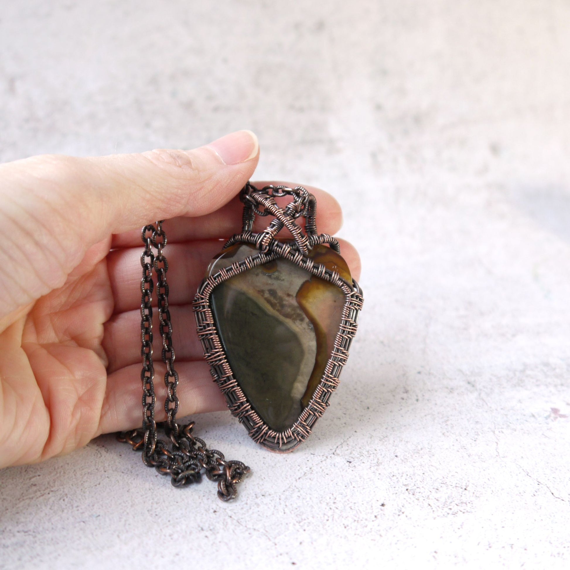 Hand holding a necklace with a large, decorative impression jasper pendant on a light background